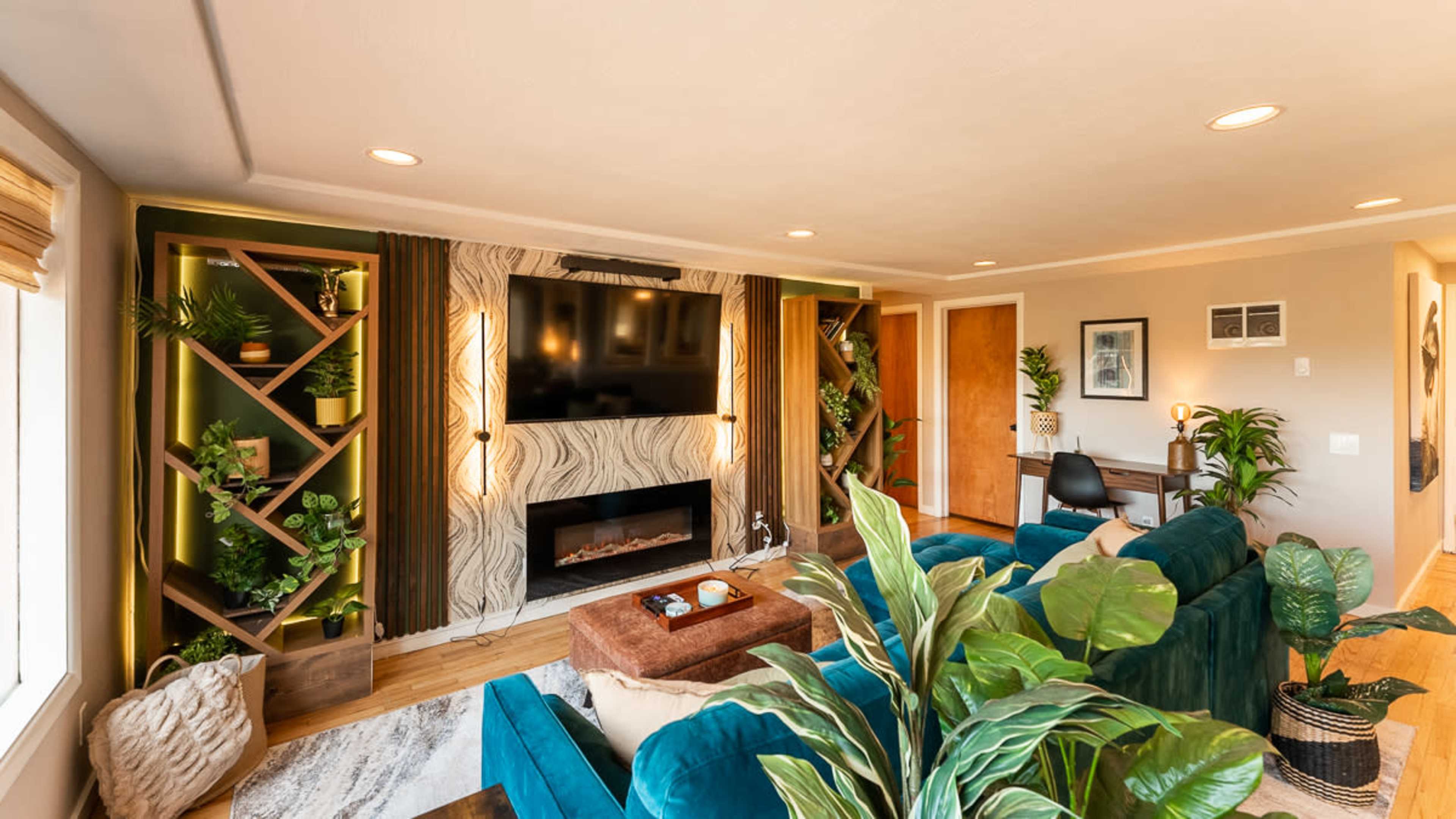 A modern living room featuring a large TV mounted on the wall, surrounded by green plants and built-in shelving, with a couch and a decorative coffee table in the center.