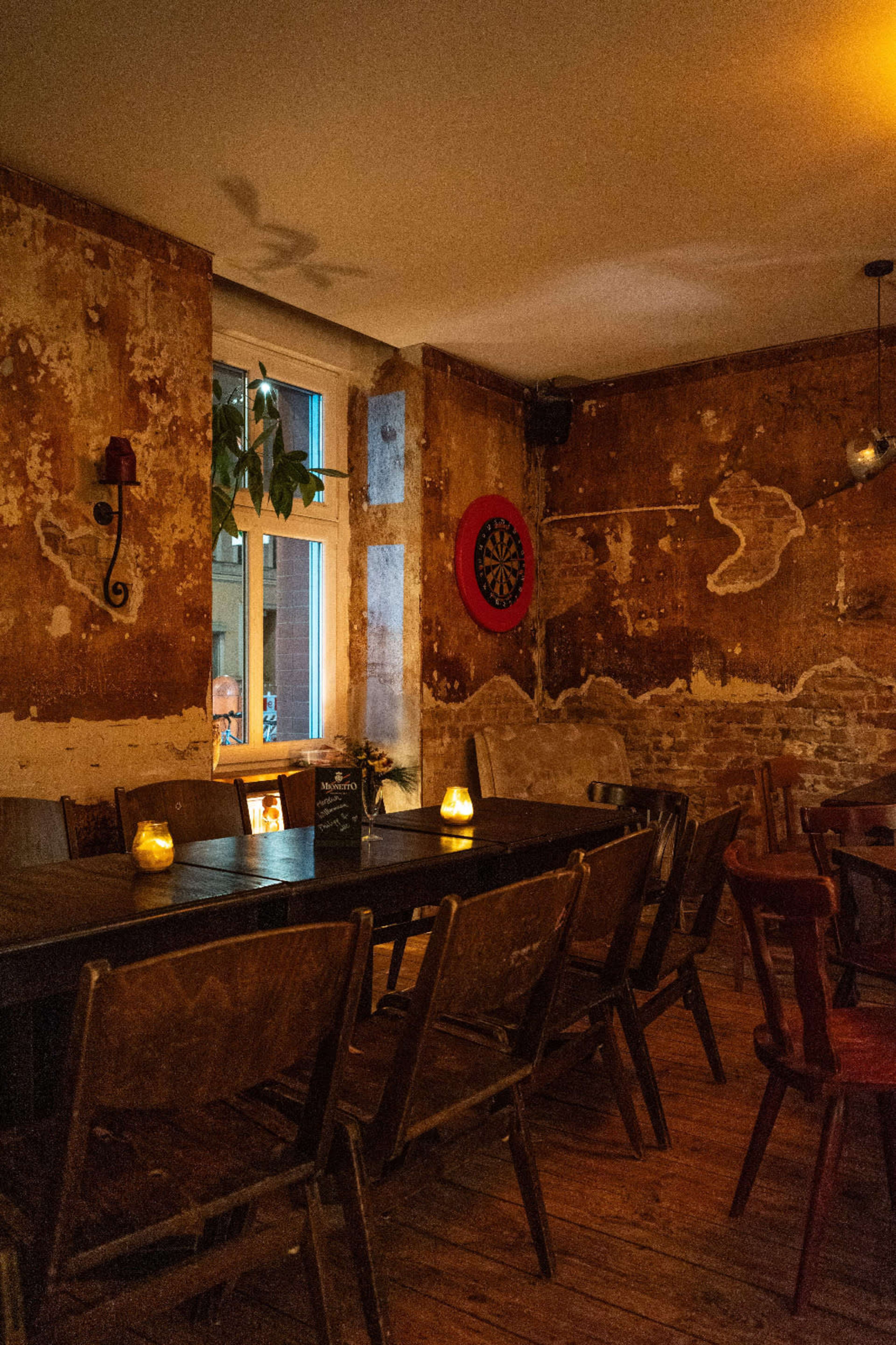 A rustic dining area features a long wooden table with several mismatched chairs, a dartboard on the wall, and candlelight illuminating the weathered walls.