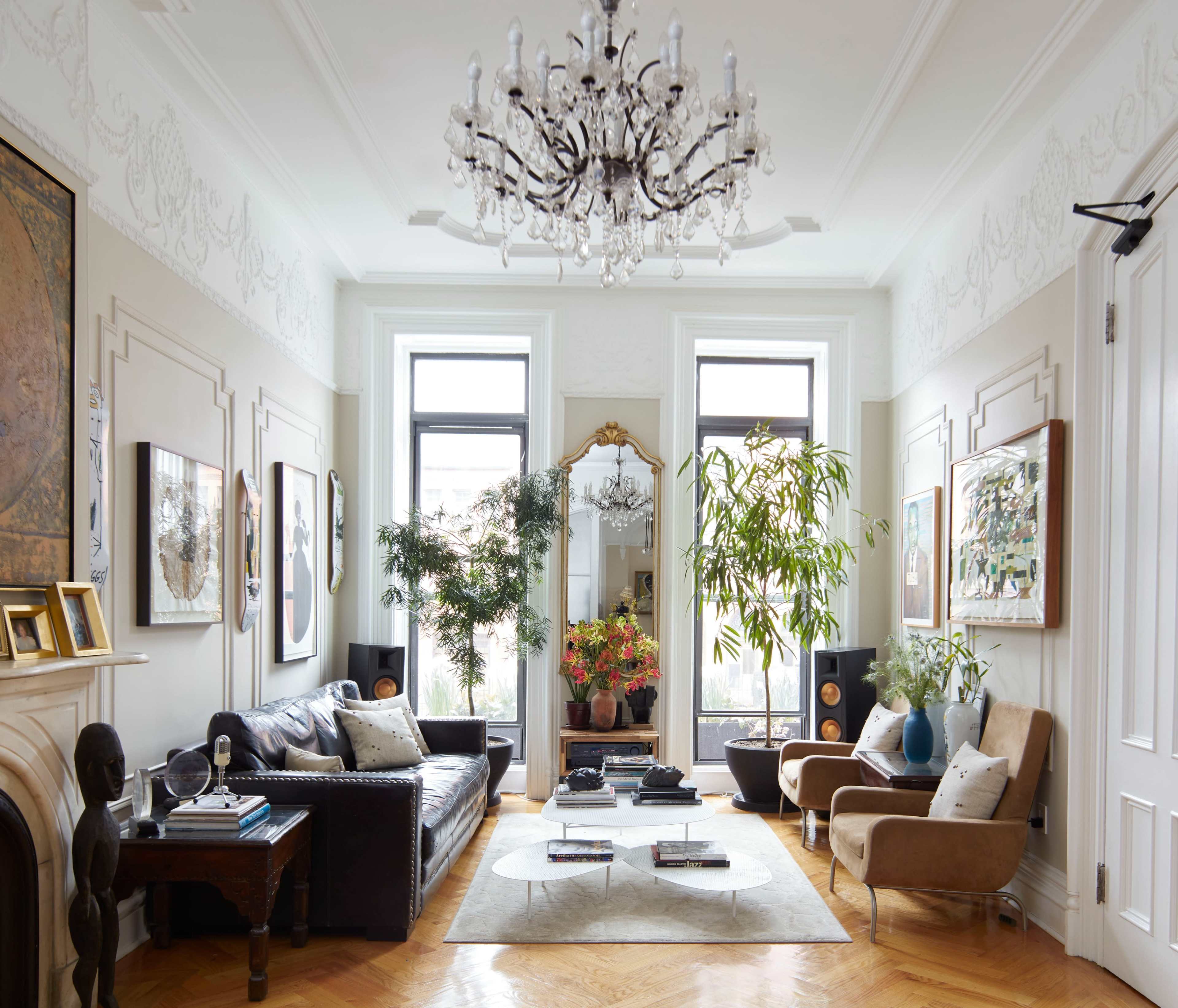 The living room features a leather sofa, two armchairs, a chandelier, large windows, and indoor plants, creating a cozy and elegant atmosphere.