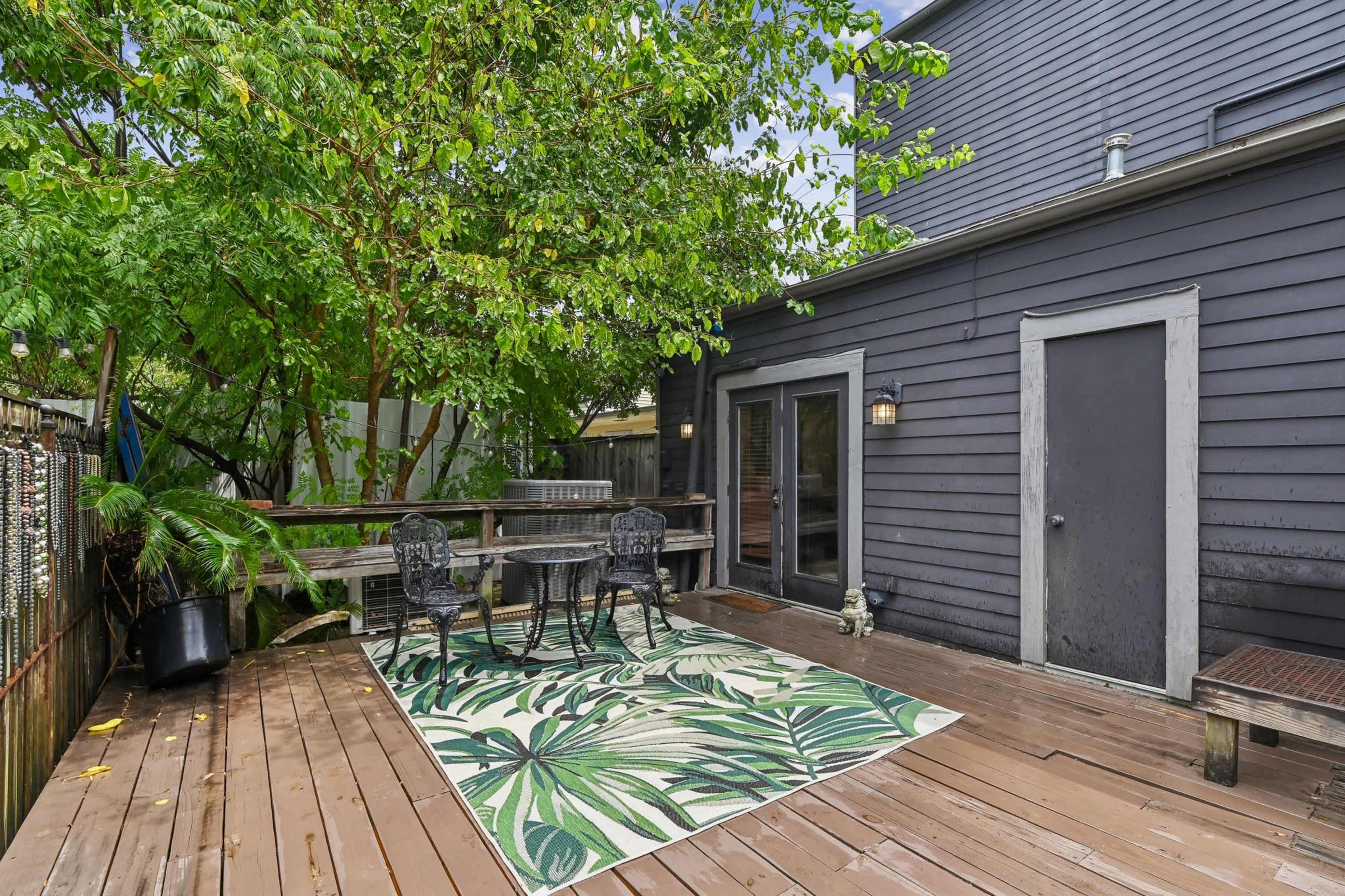 The image shows a wooden deck featuring a green and white patterned rug with a table and chairs set against a backdrop of a dark exterior wall and lush greenery.