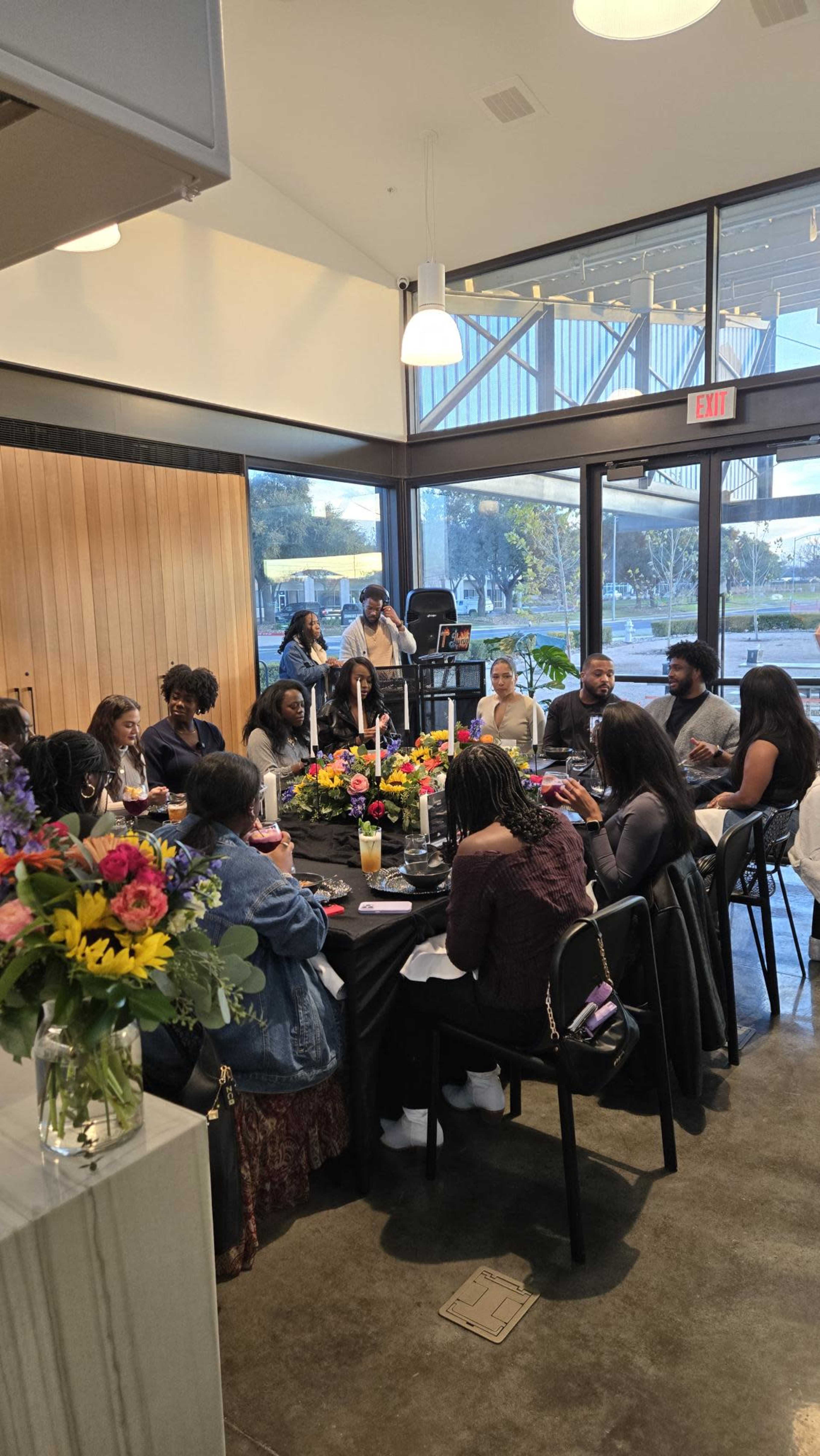 A group of people seated around a table covered with flowers, food, and drinks is engaged in conversation in a well-lit, modern indoor setting.