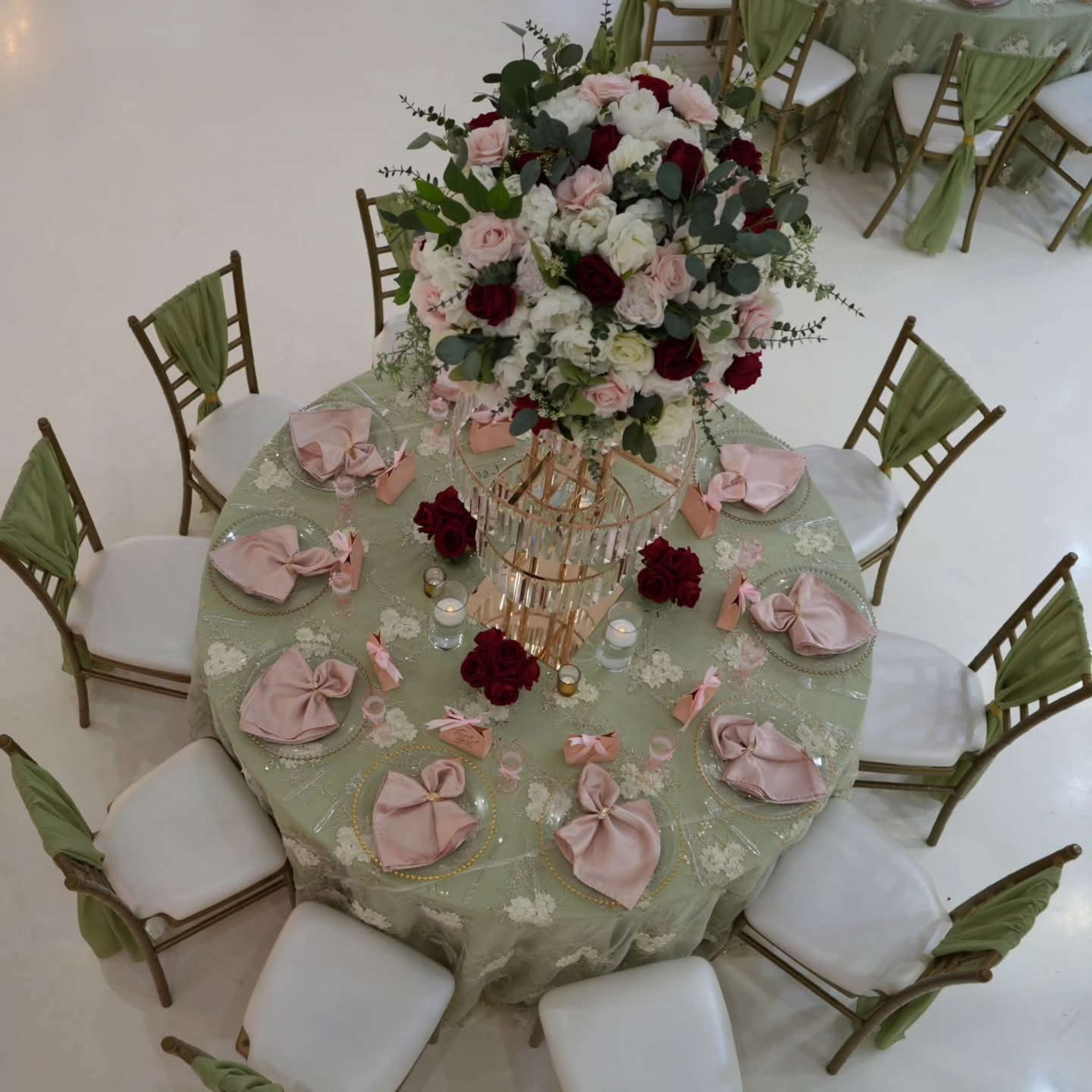 A large round table is elegantly set with a floral centerpiece, pink napkins, and decorative accents, surrounded by gold-framed chairs.