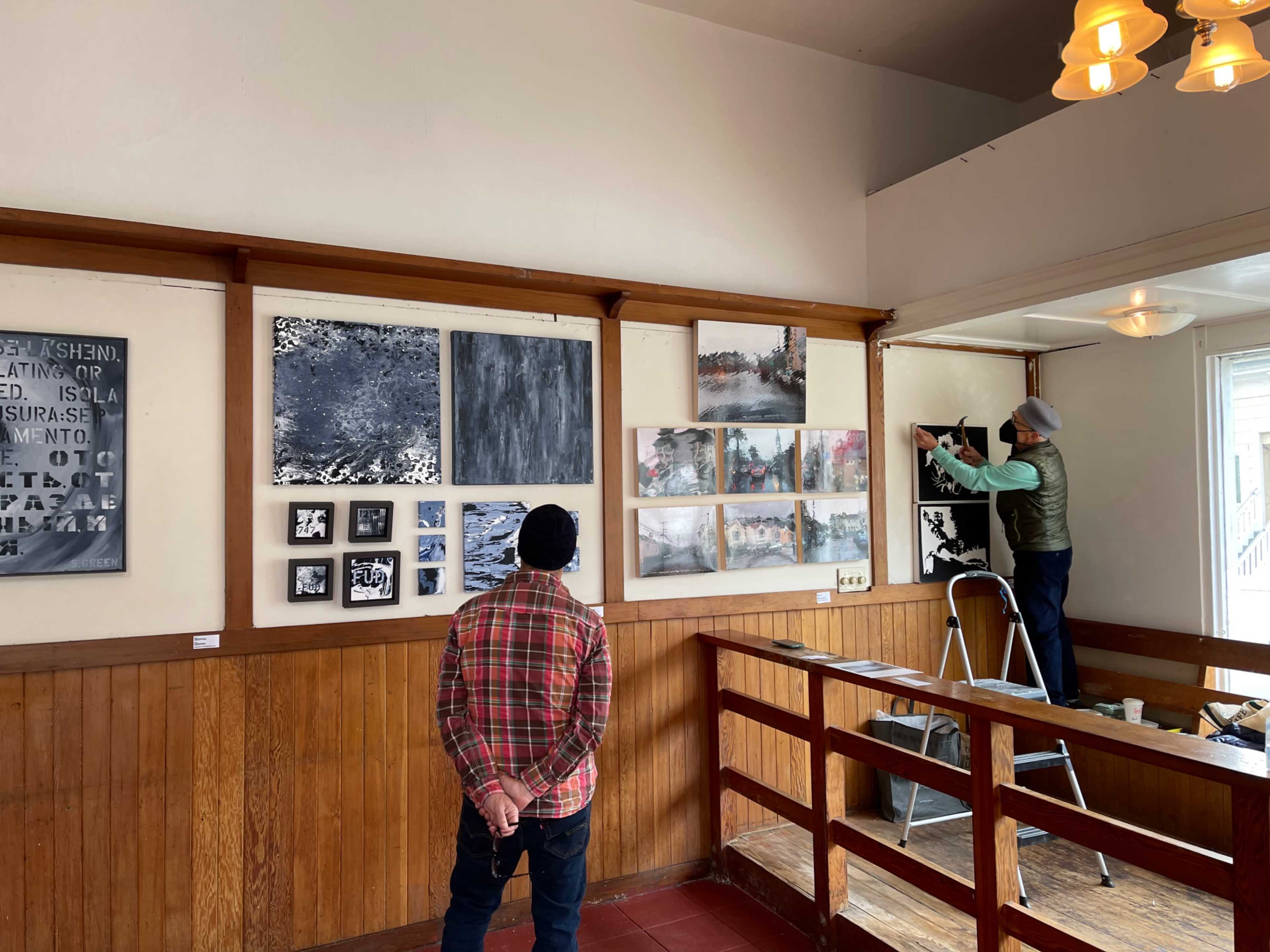 Two people are inside an art gallery; one man stands observing the artwork on the wall while another man is hanging a painting.