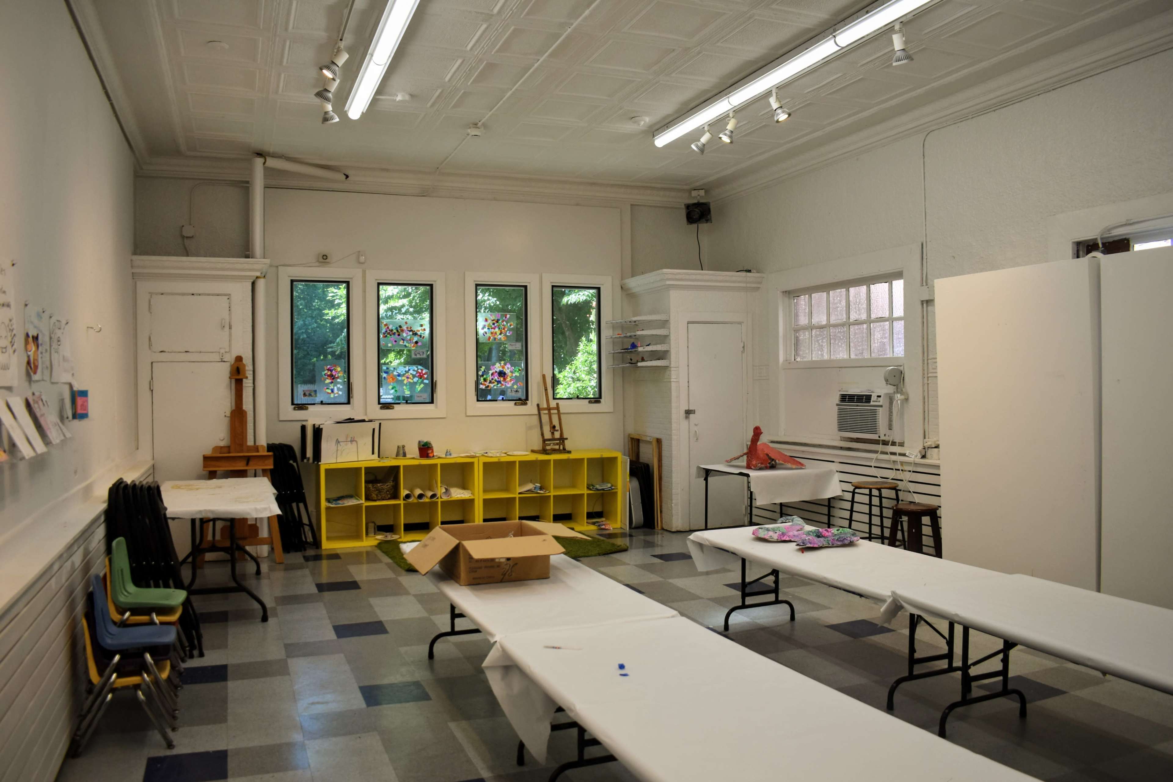 The image shows a spacious art classroom with tables covered in white cloths, colorful stained glass windows, and yellow storage shelves along the wall.