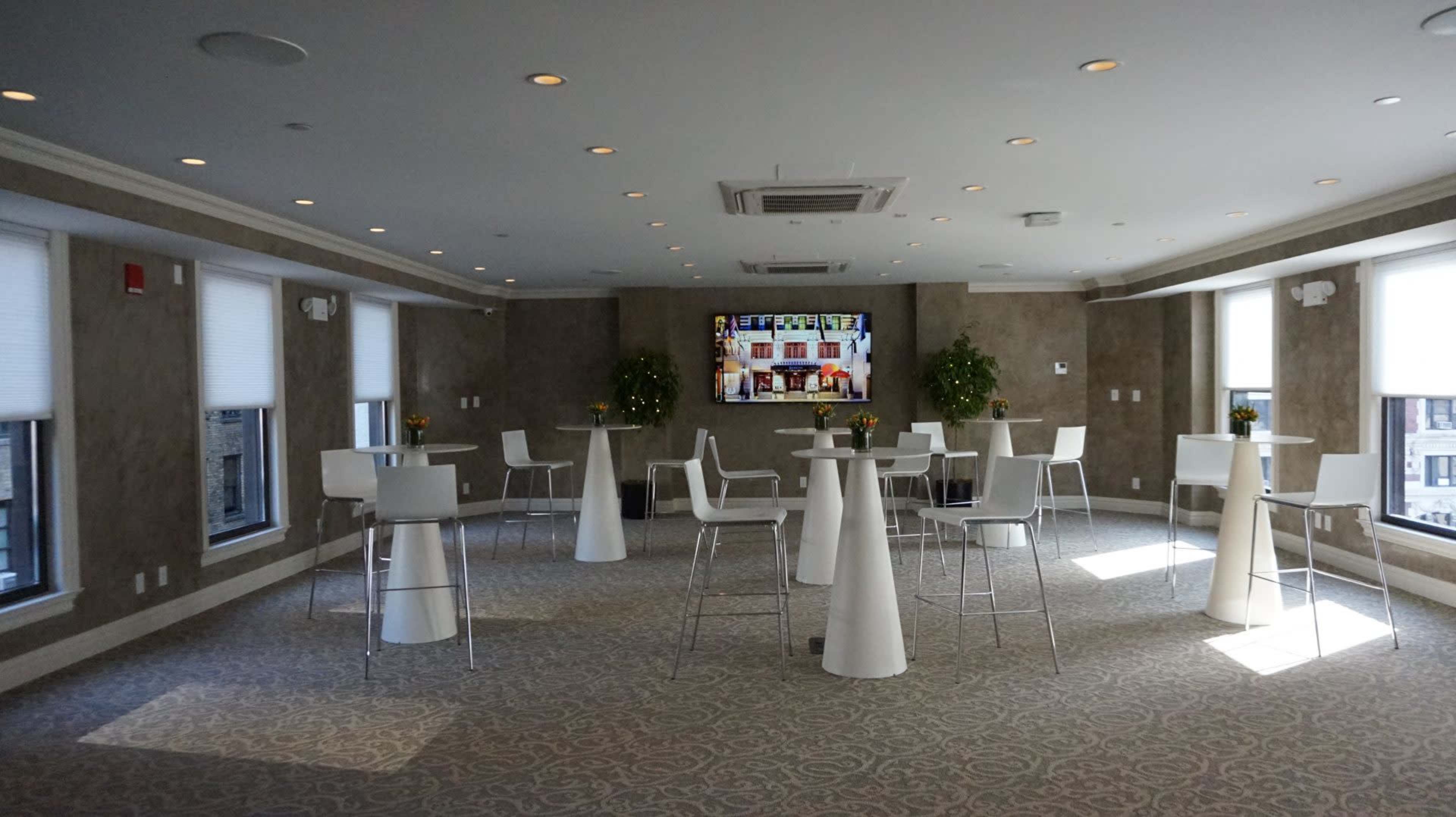 A spacious event room features tall white tables and chairs, with a large television screen and potted plants along the windows.