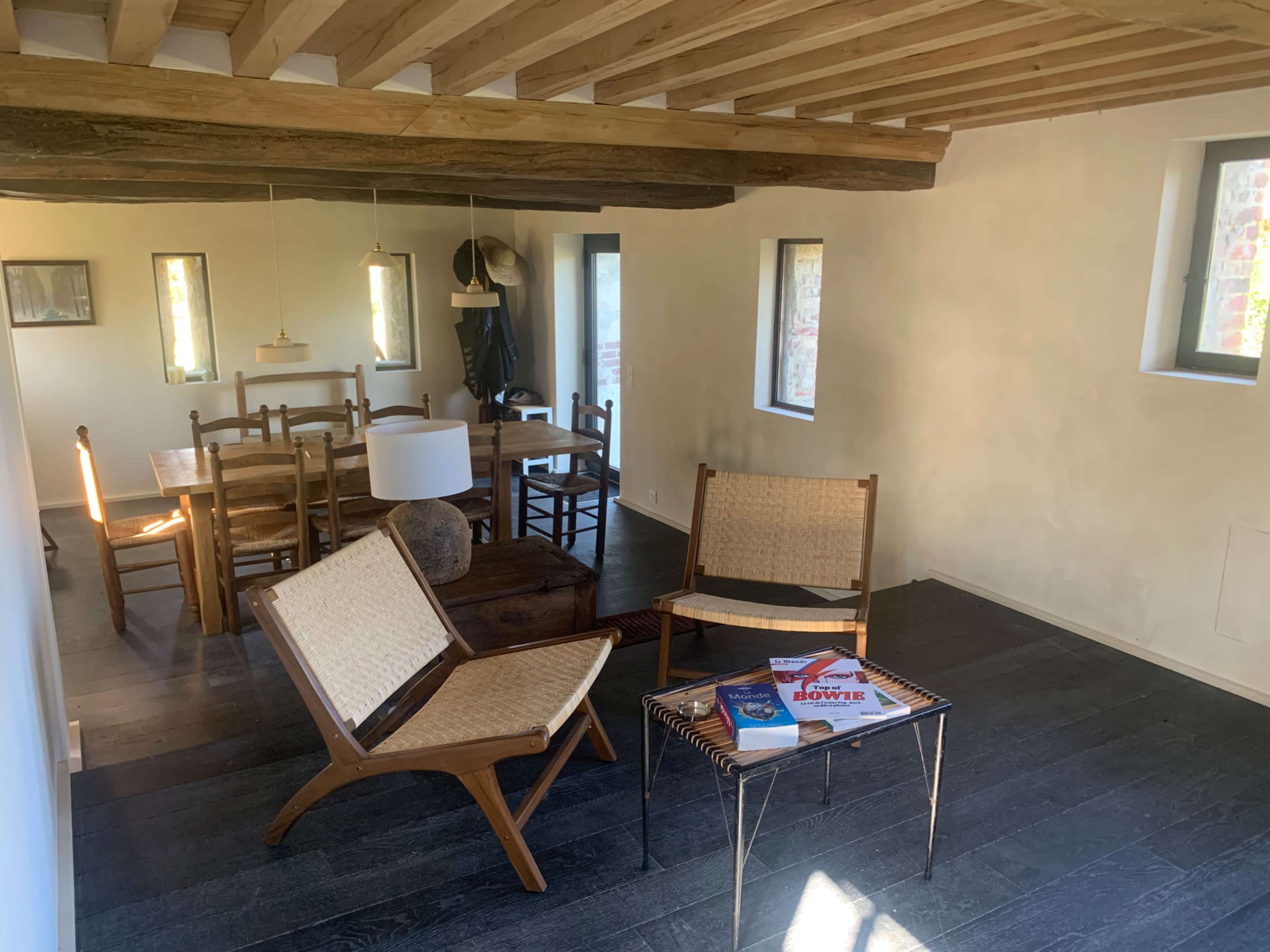 The image shows a lightly furnished room with wooden beams, featuring two chairs, a coffee table with books, and a dining area with a wooden table and chairs.