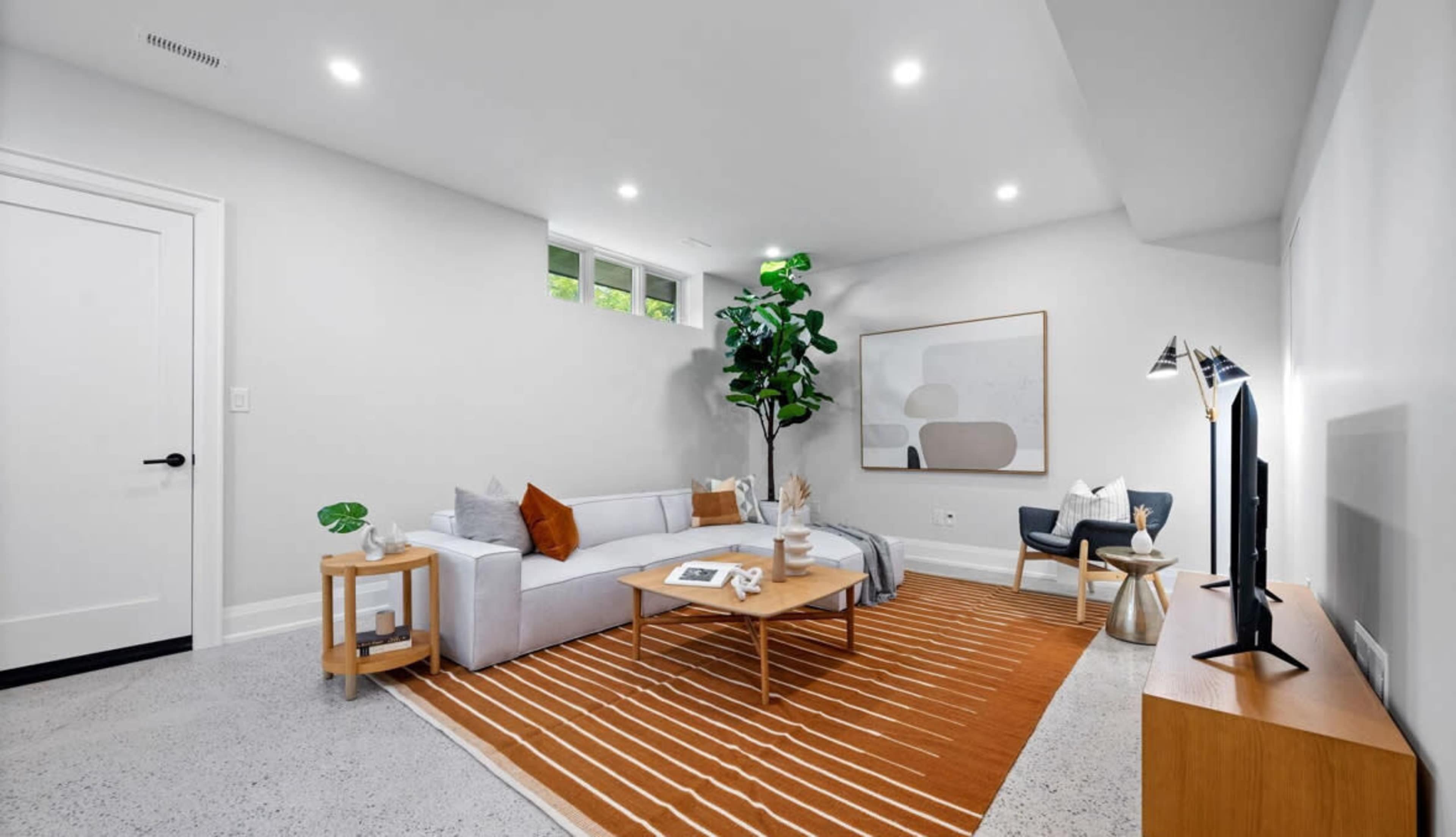 The image shows a modern living room featuring a light gray sofa, a wooden coffee table, a large area rug with orange stripes, and a television stand against a white wall.