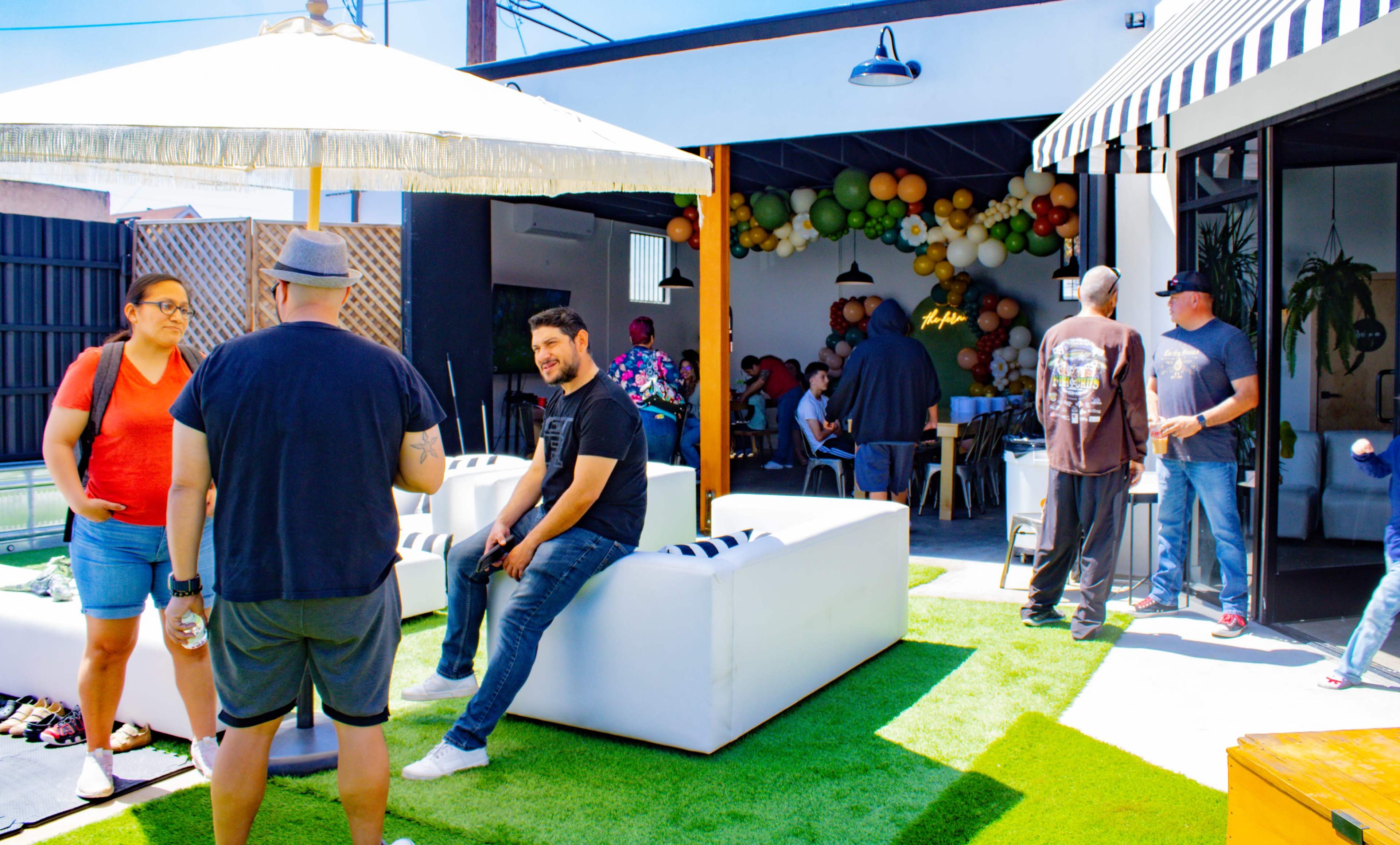 A group of people socializes in a landscaped outdoor space featuring white seating, a large umbrella, and colorful decorations.