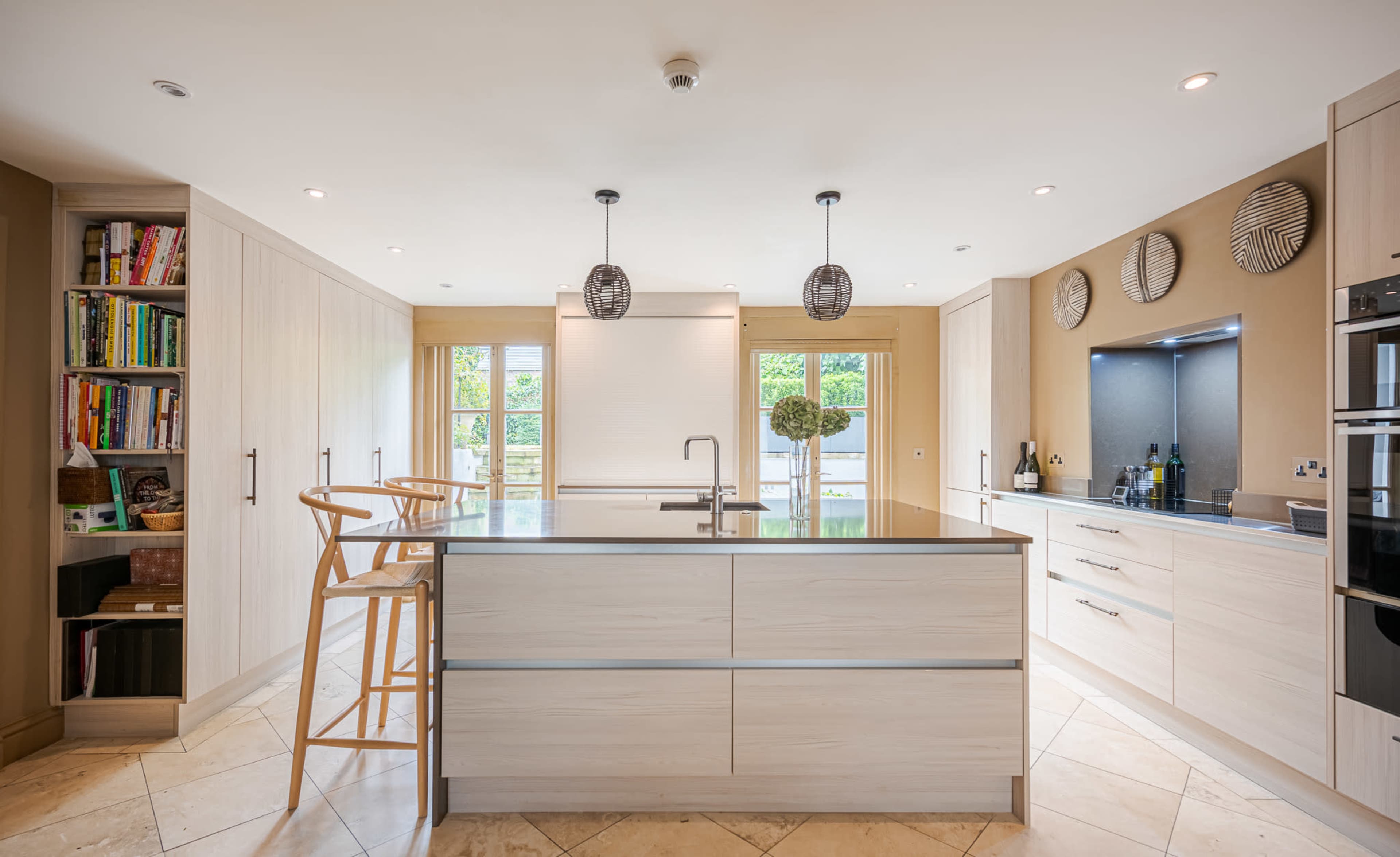 The image shows a modern kitchen featuring a central island, light wood cabinetry, and large windows allowing natural light to enter the space.