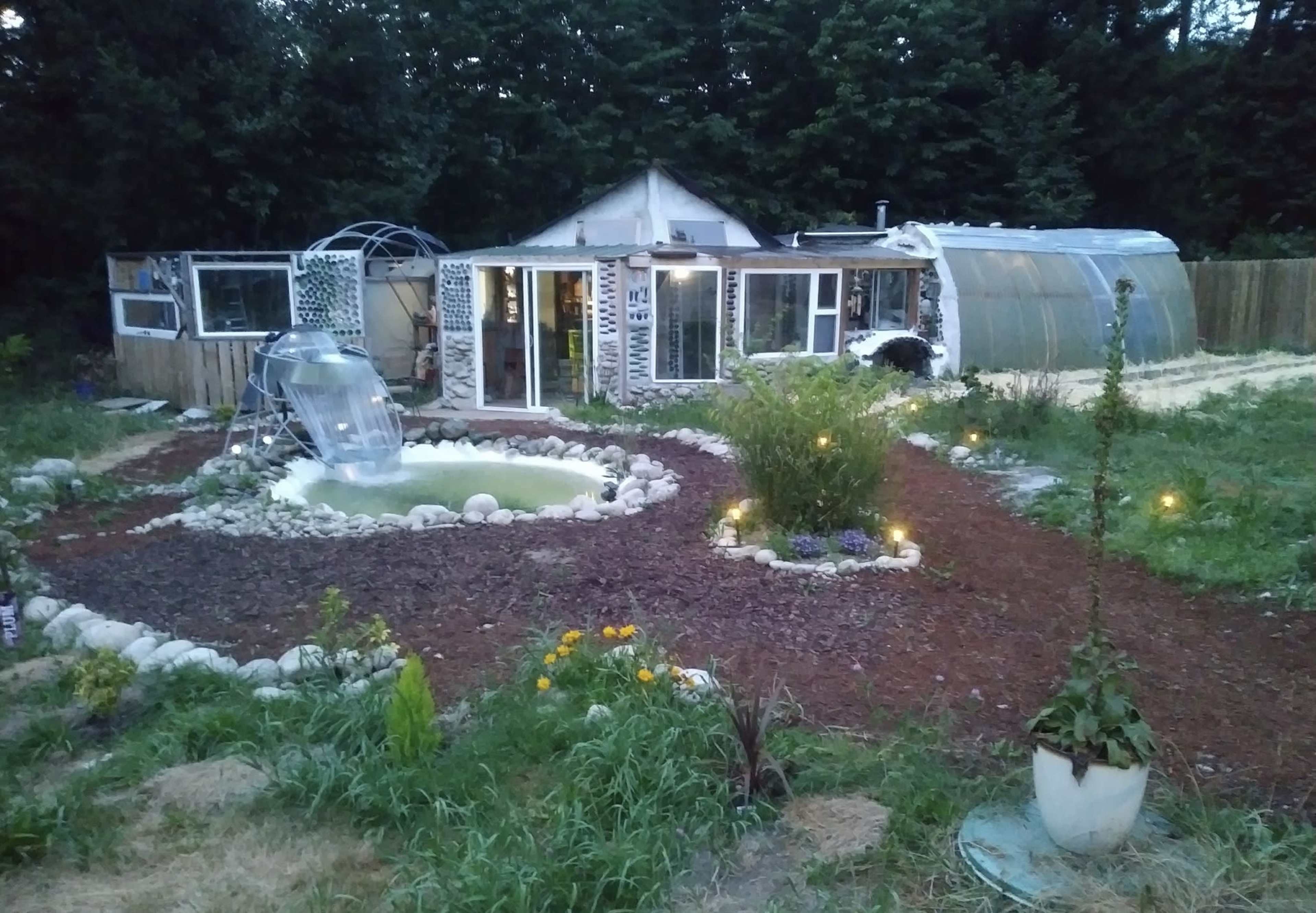A garden with a small pond and a water slide, surrounded by a rocky path, leading to a greenhouse and a house in the background.