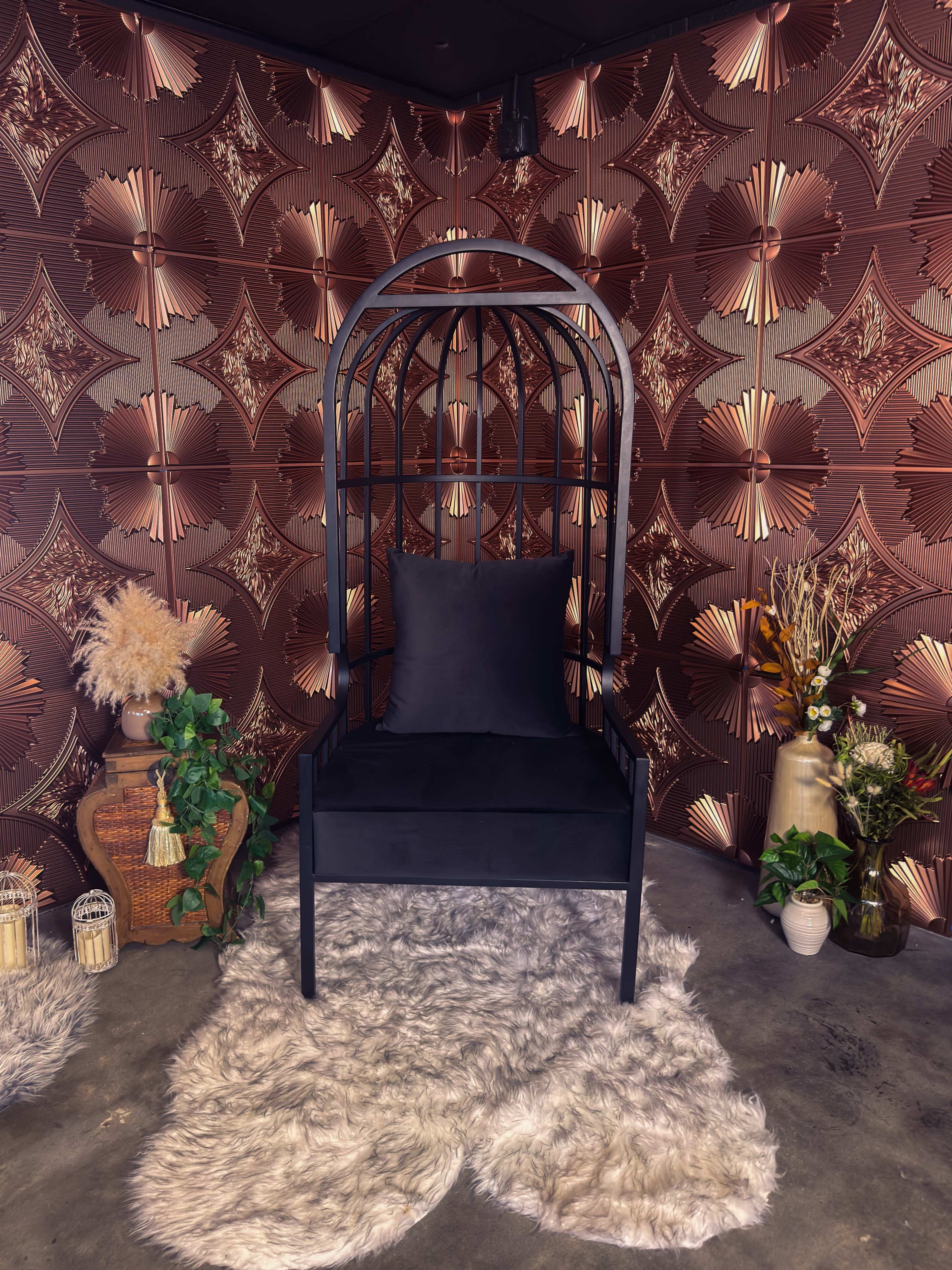 The image shows a black chair with an arching frame placed on a fluffy white rug against a patterned brown wall adorned with geometric designs and decorative plants.