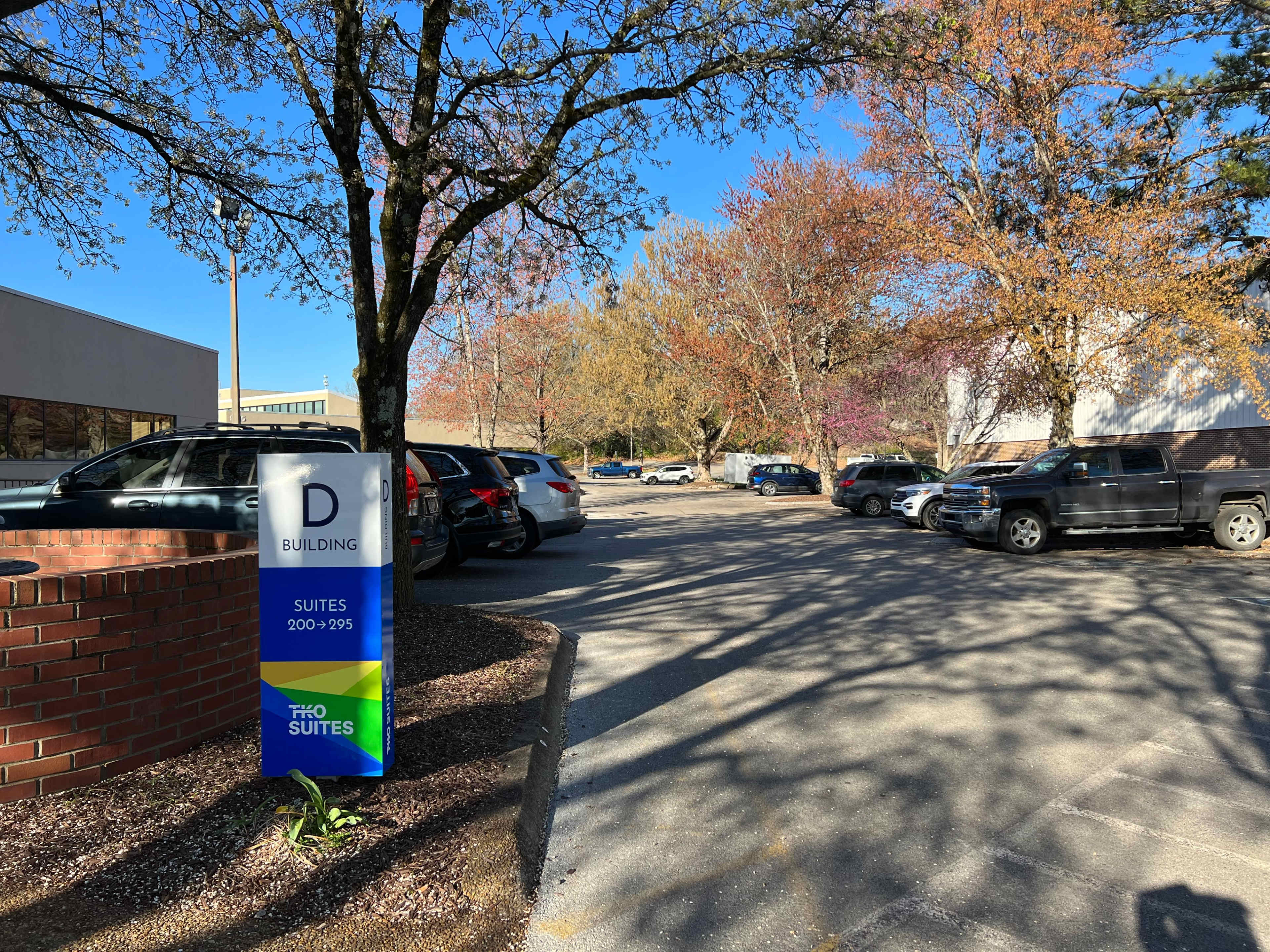 A sign indicating "D BUILDING" with suites numbered 900 to 295 stands at the entrance of a parking lot lined with vehicles and trees displaying autumn foliage.