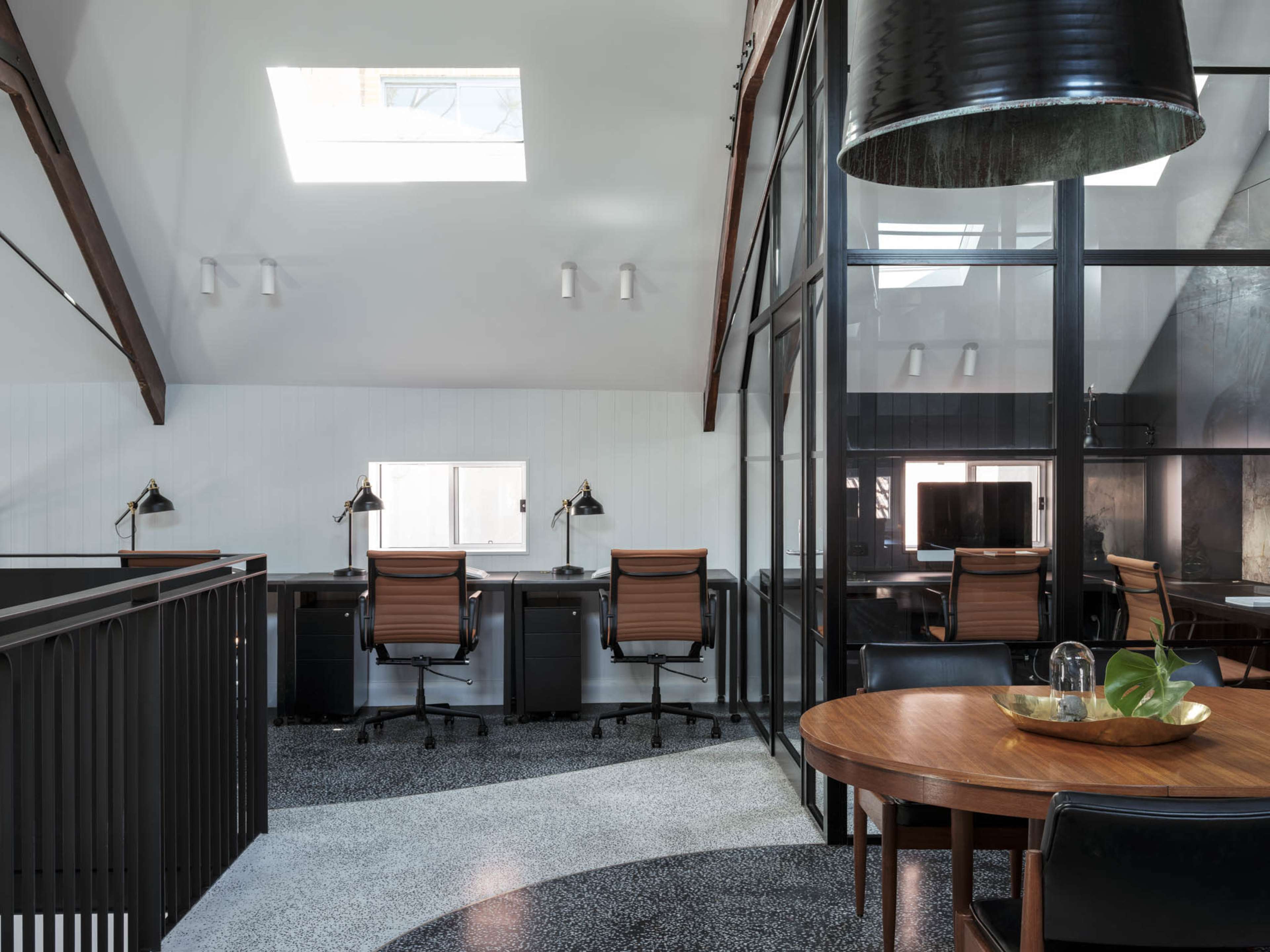 The image shows a modern office space featuring a mix of workstations with desk lamps, a large round table, and large windows allowing natural light.