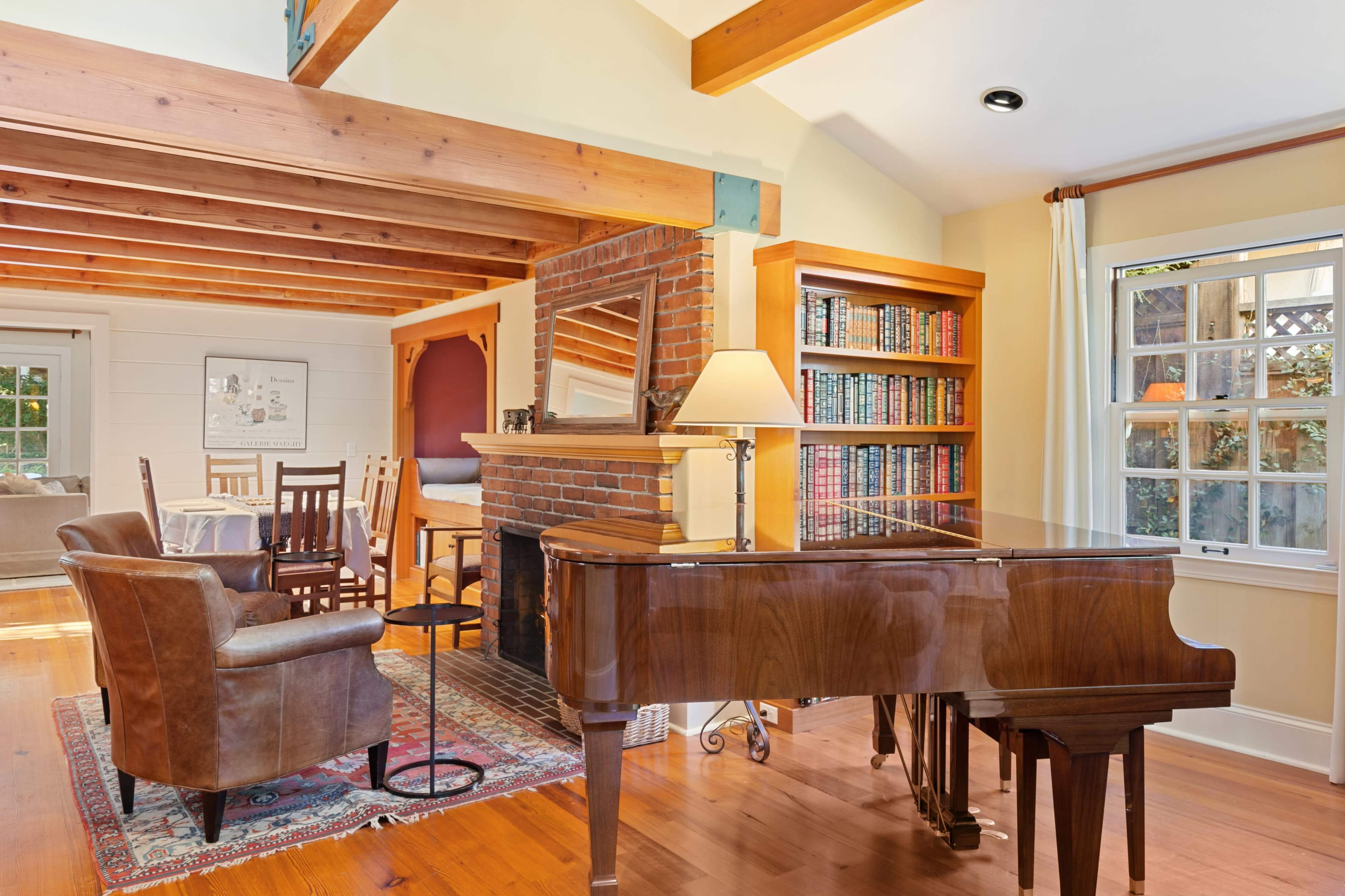 A spacious, well-lit room featuring a grand piano, a seating area with chairs, a dining table, and a bookshelf lined with books.