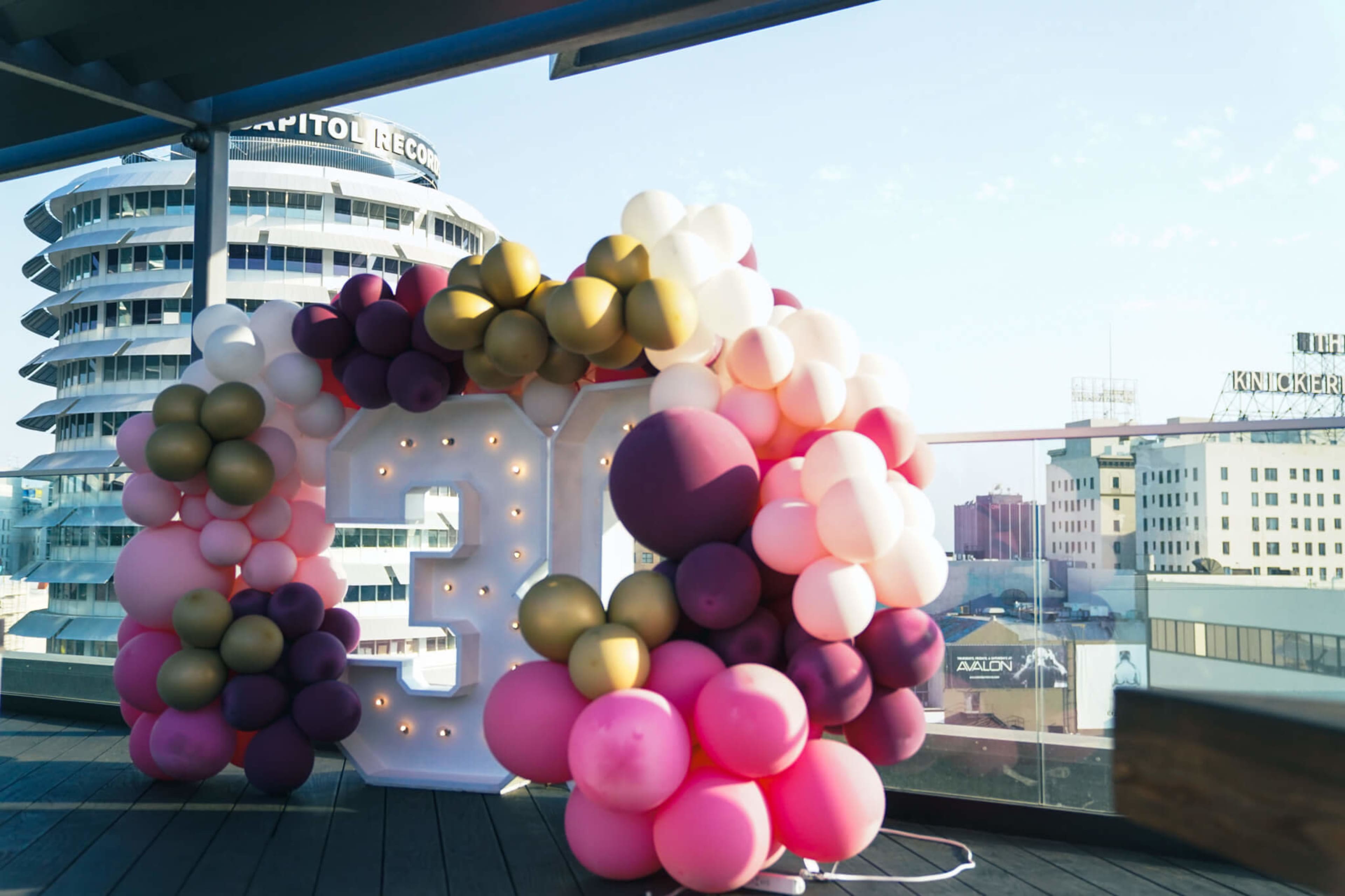 A large "30" made of illuminated numbers is surrounded by colorful balloons on a rooftop terrace with a city skyline in the background.