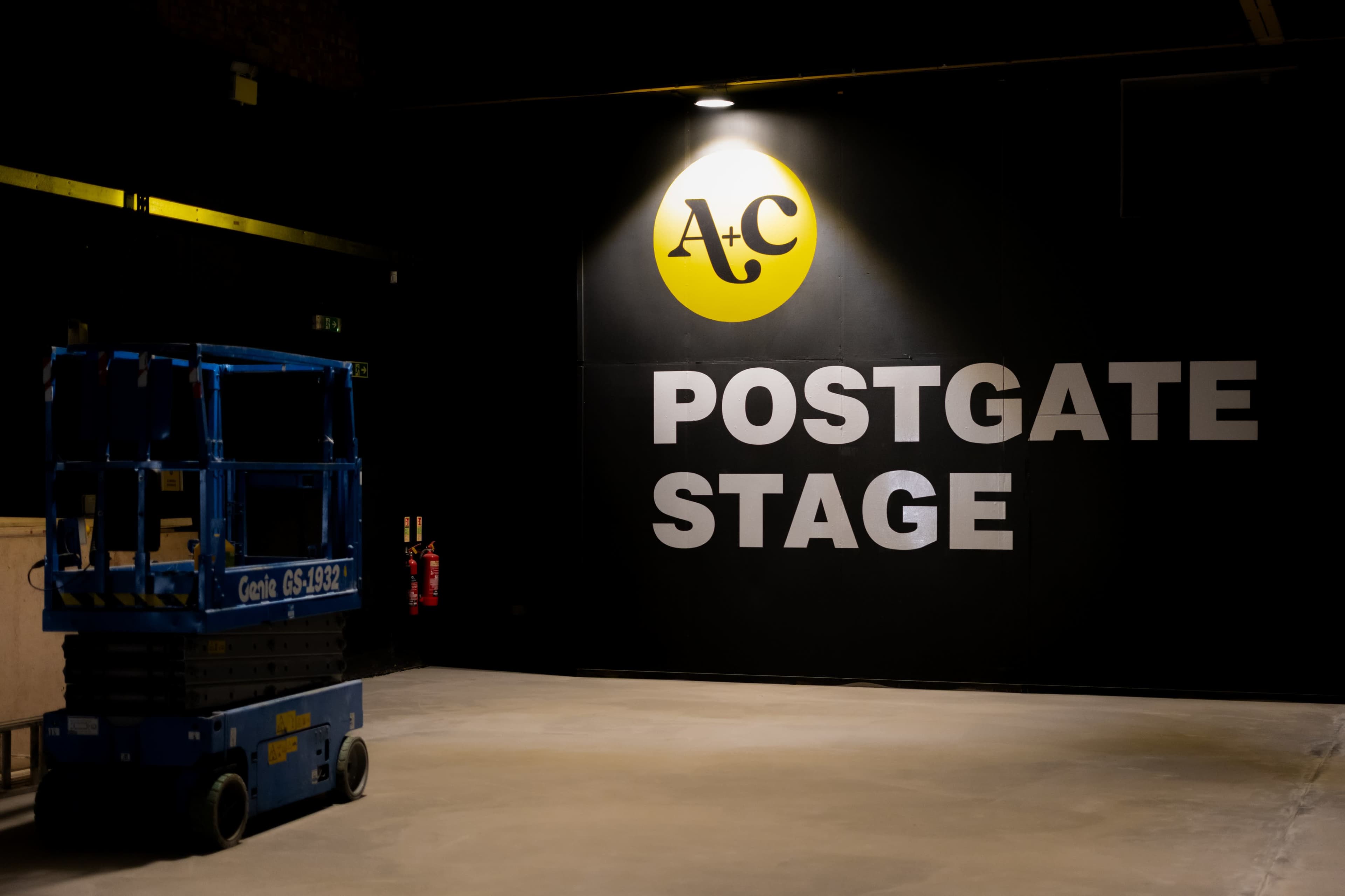 The image shows an industrial space with a large wall displaying the words "POSTGATE STAGE" alongside a yellow logo, and a blue scissor lift is positioned in the foreground.