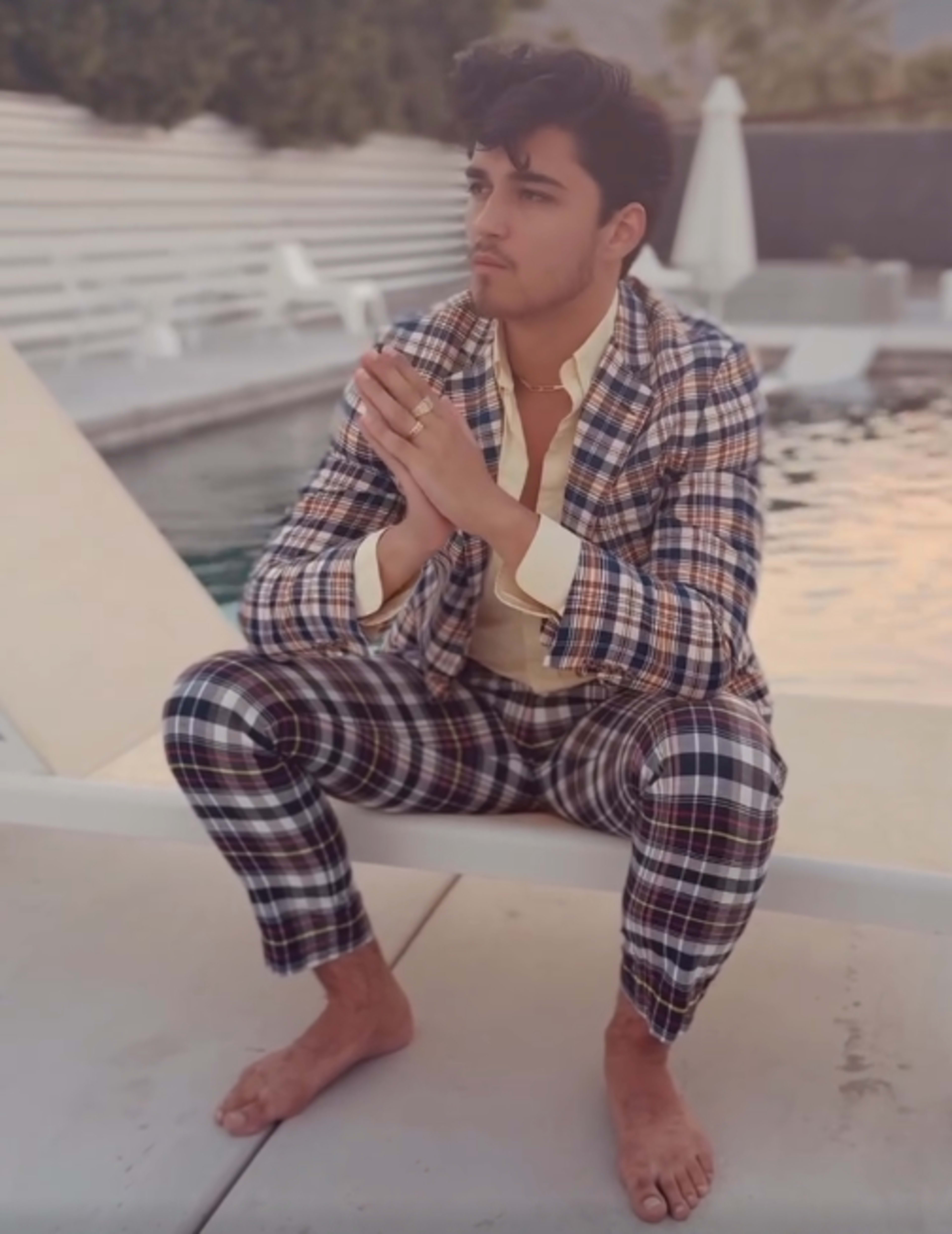 A man in a plaid suit sits by a swimming pool, barefoot, with his hands clasped in front of him.