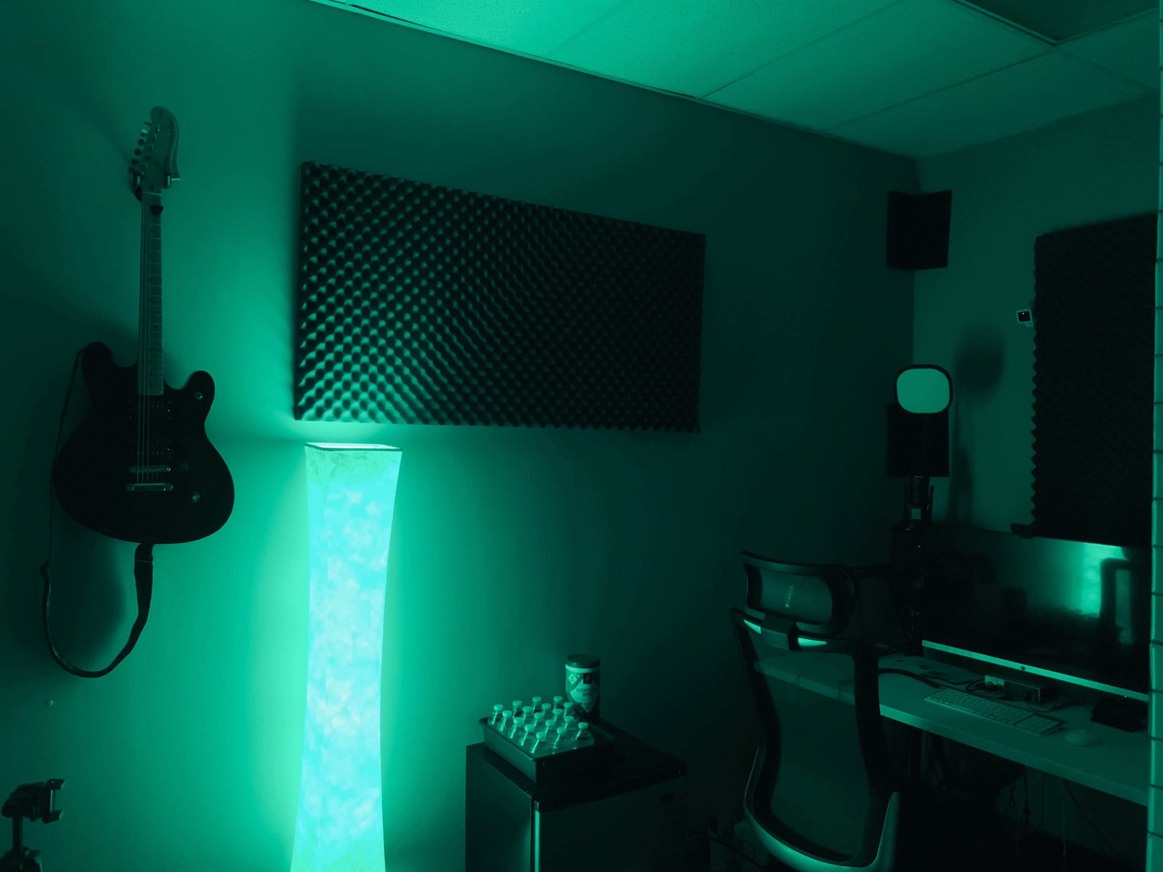 A dimly lit room with green walls, featuring a guitar hanging on the wall, a glowing floor lamp, and a desk with a computer and office chair.