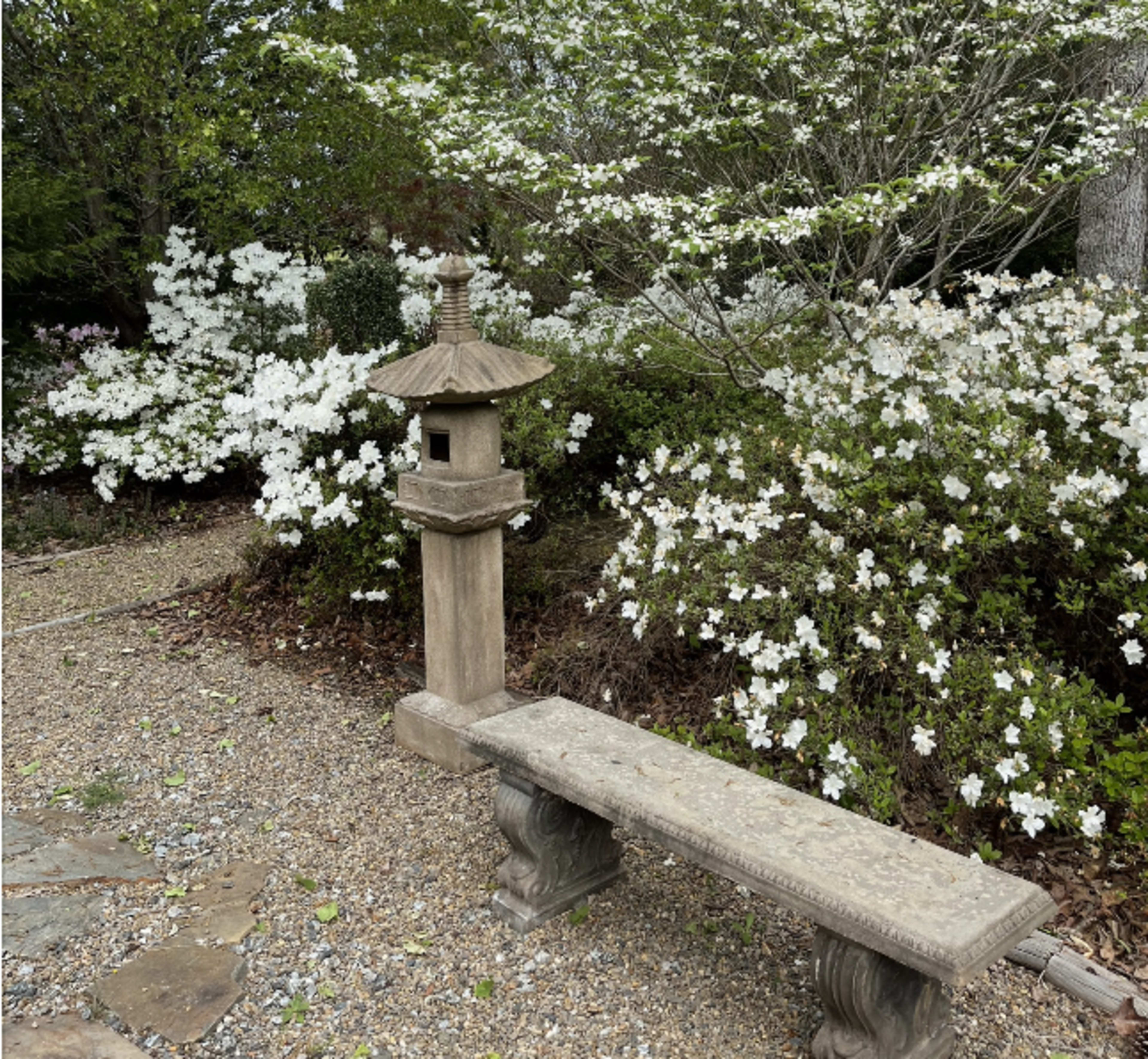 A stone lantern and a concrete bench are situated near flowering white azaleas in a landscaped garden.
