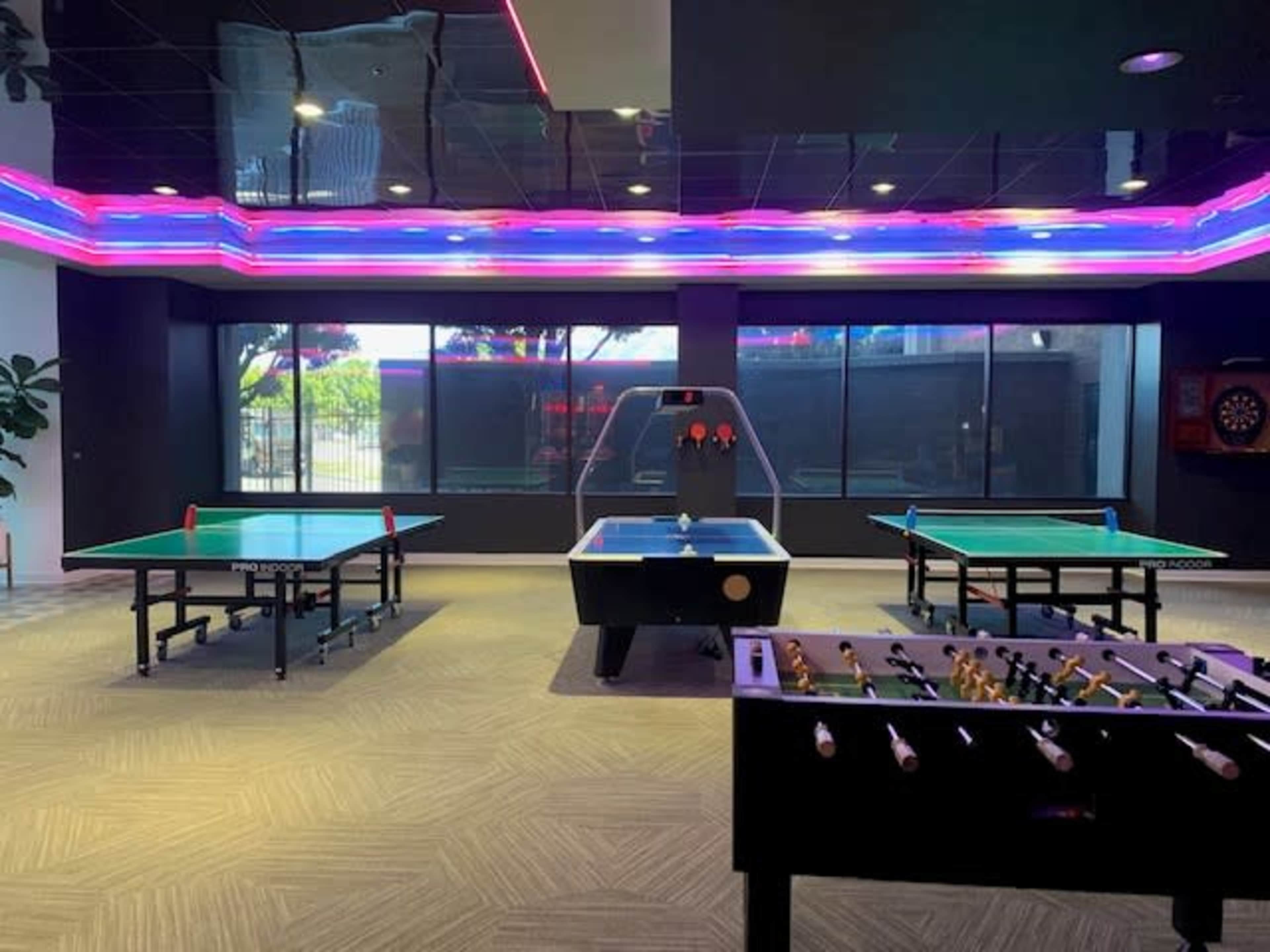 A recreational room featuring multiple table games, including ping pong tables, a pool table, and a foosball table, all under colorful neon lighting.