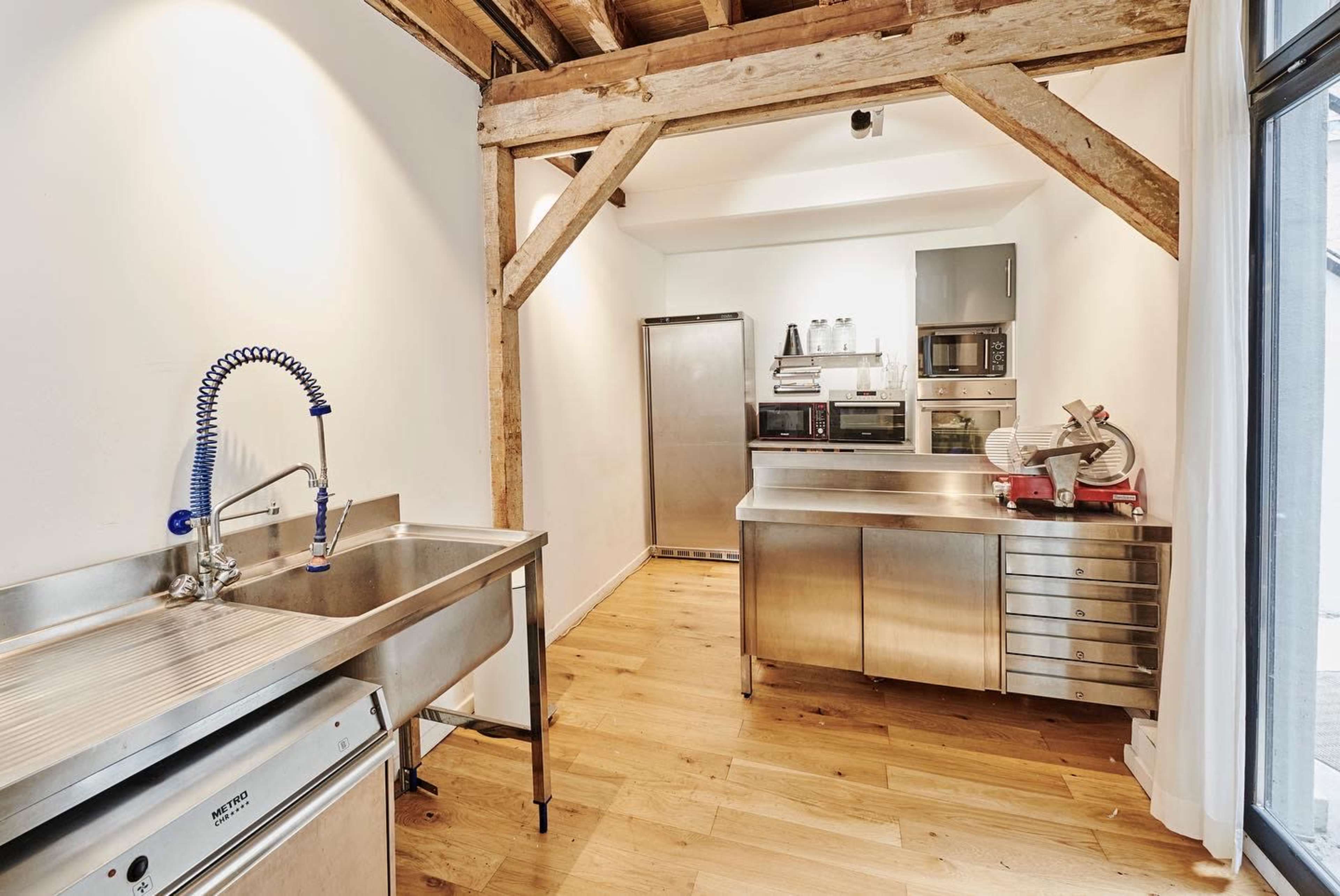 A modern kitchen with stainless steel appliances, a workbench, and a large sink, set against exposed wooden beams.