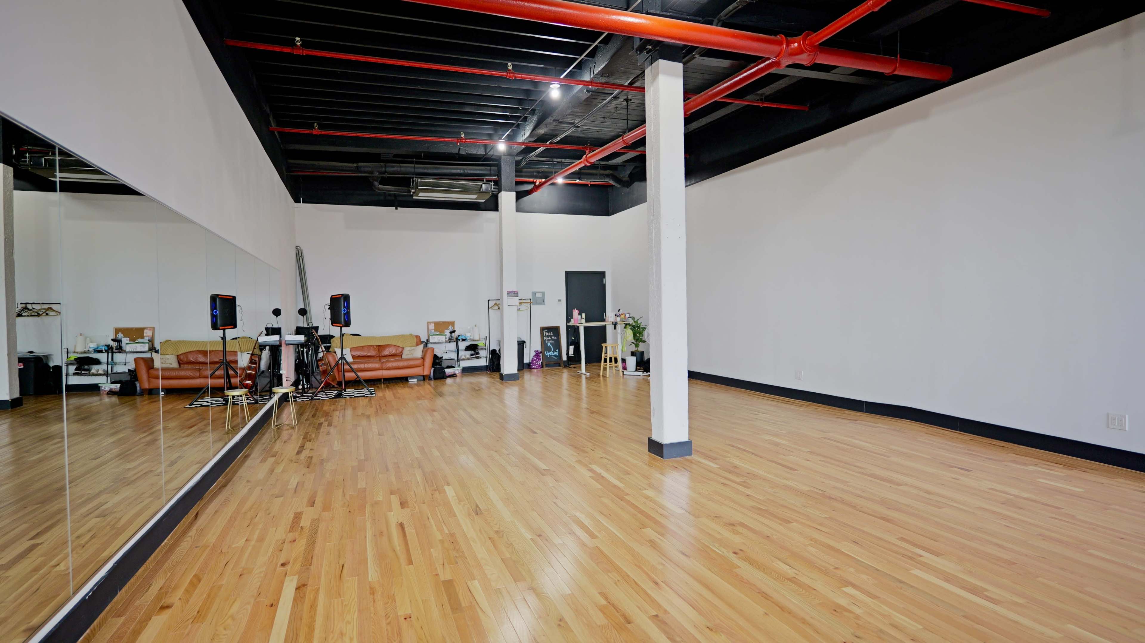 The image shows a spacious dance studio with a wooden floor, large mirrors on one wall, and minimalist furnishings, including a couch and audio equipment.