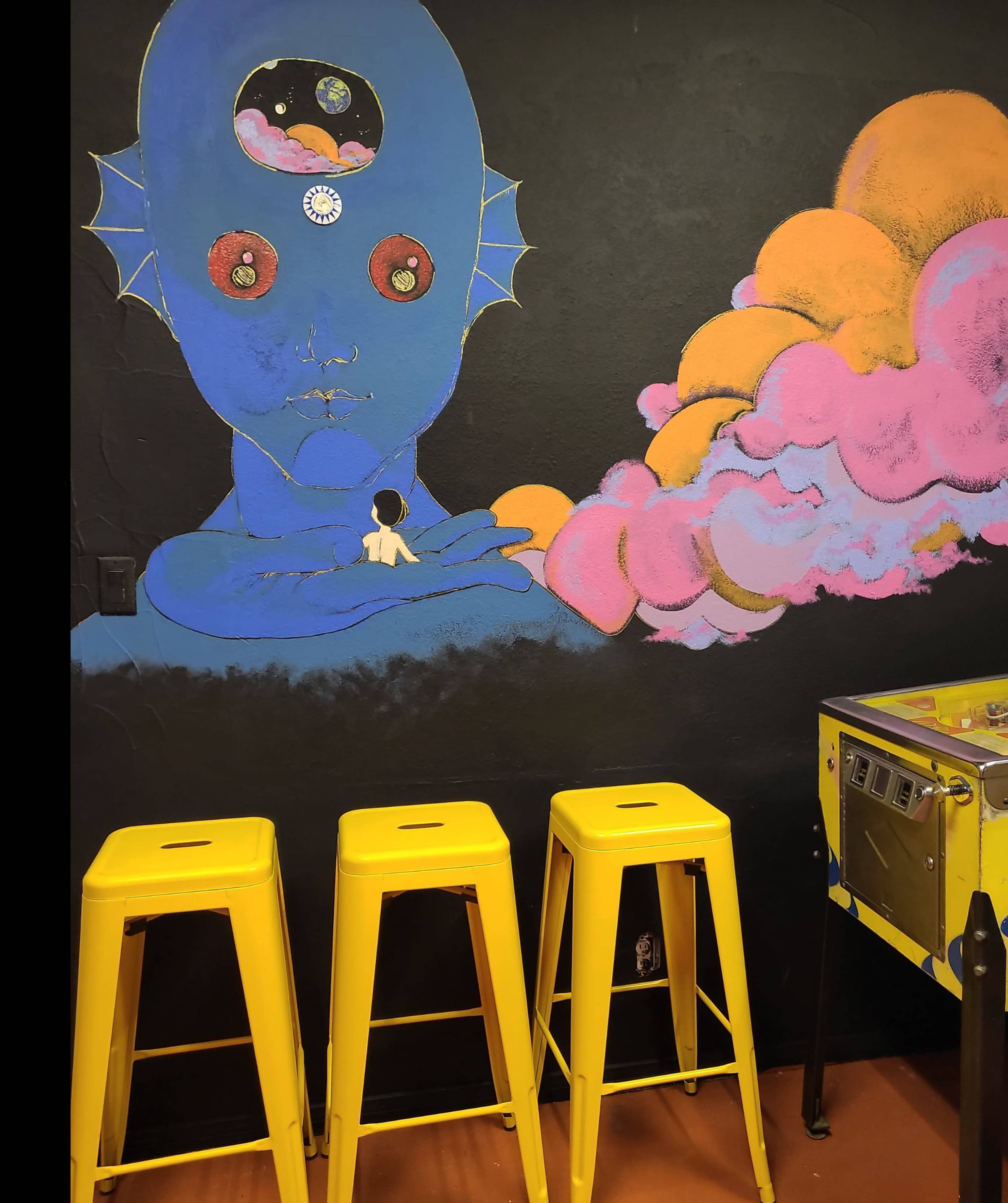 A mural of a blue-skinned figure with a cosmic head is painted on a black wall beside three yellow stools and a pinball machine.