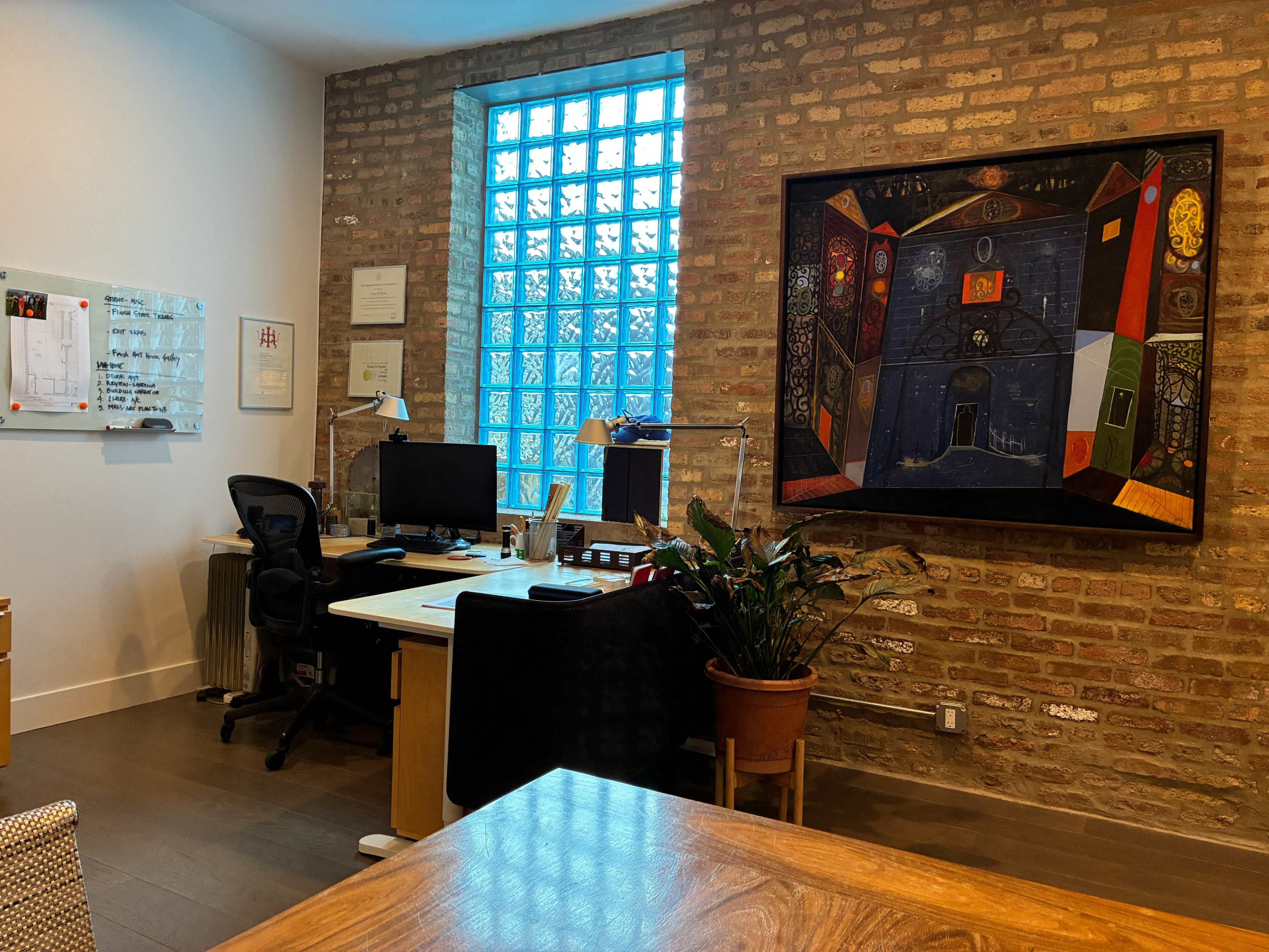 The image shows a modern office space featuring a desk with a computer, a large wall-mounted artwork, and a potted plant beside a blue window.