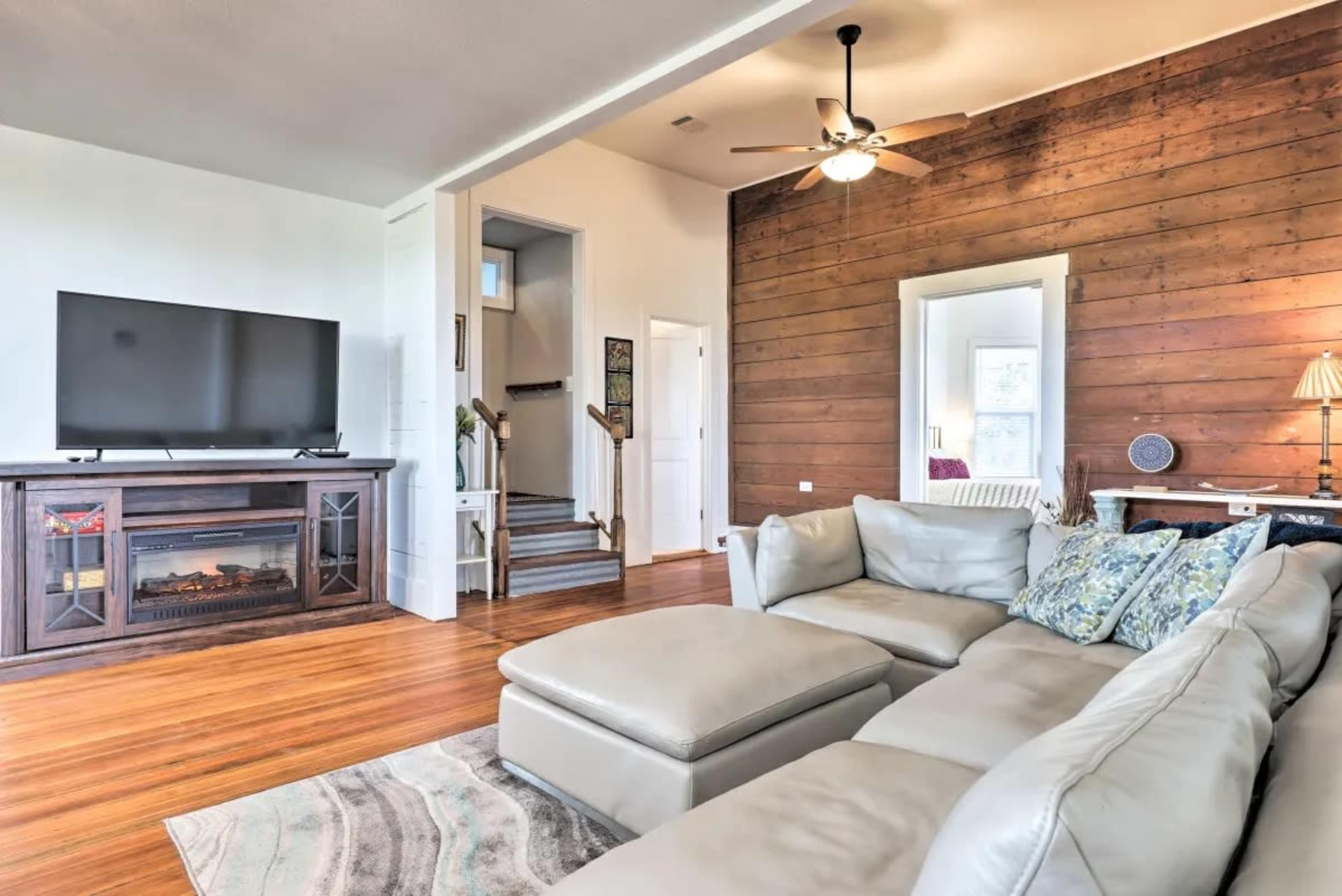 The living room features a sectional sofa, a flat-screen TV on a wooden stand, and a staircase leading to an upper level, with a mirror reflecting the space.
