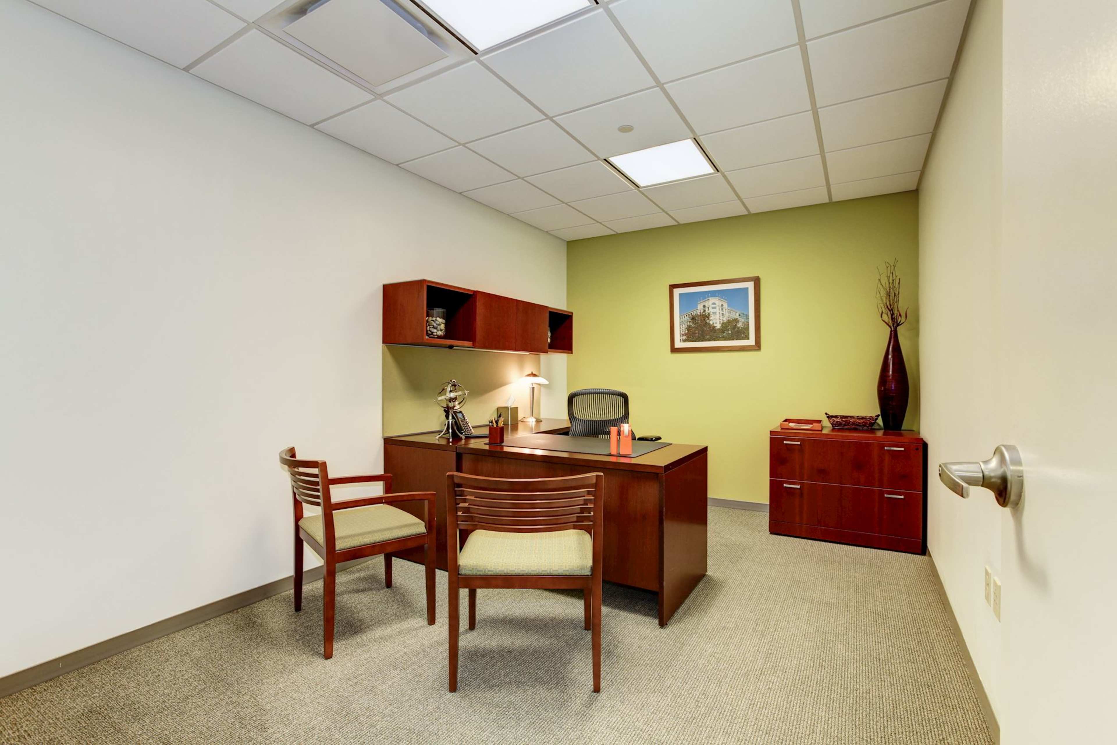 The image shows a sparse office space featuring a wooden desk, two chairs, and a green accent wall with a framed picture.
