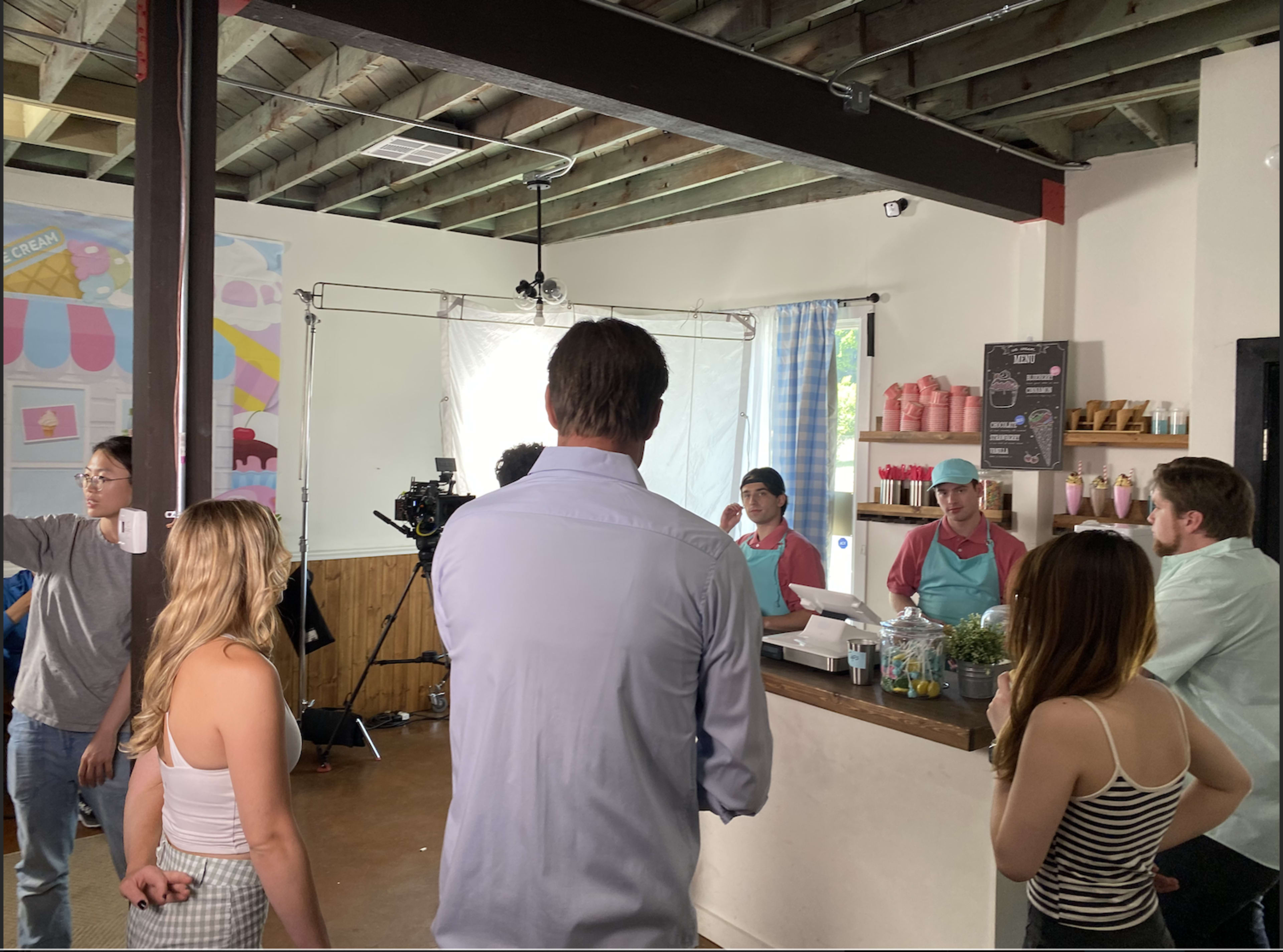 A film crew records a scene at a brightly decorated ice cream shop with actors interacting at the service counter.