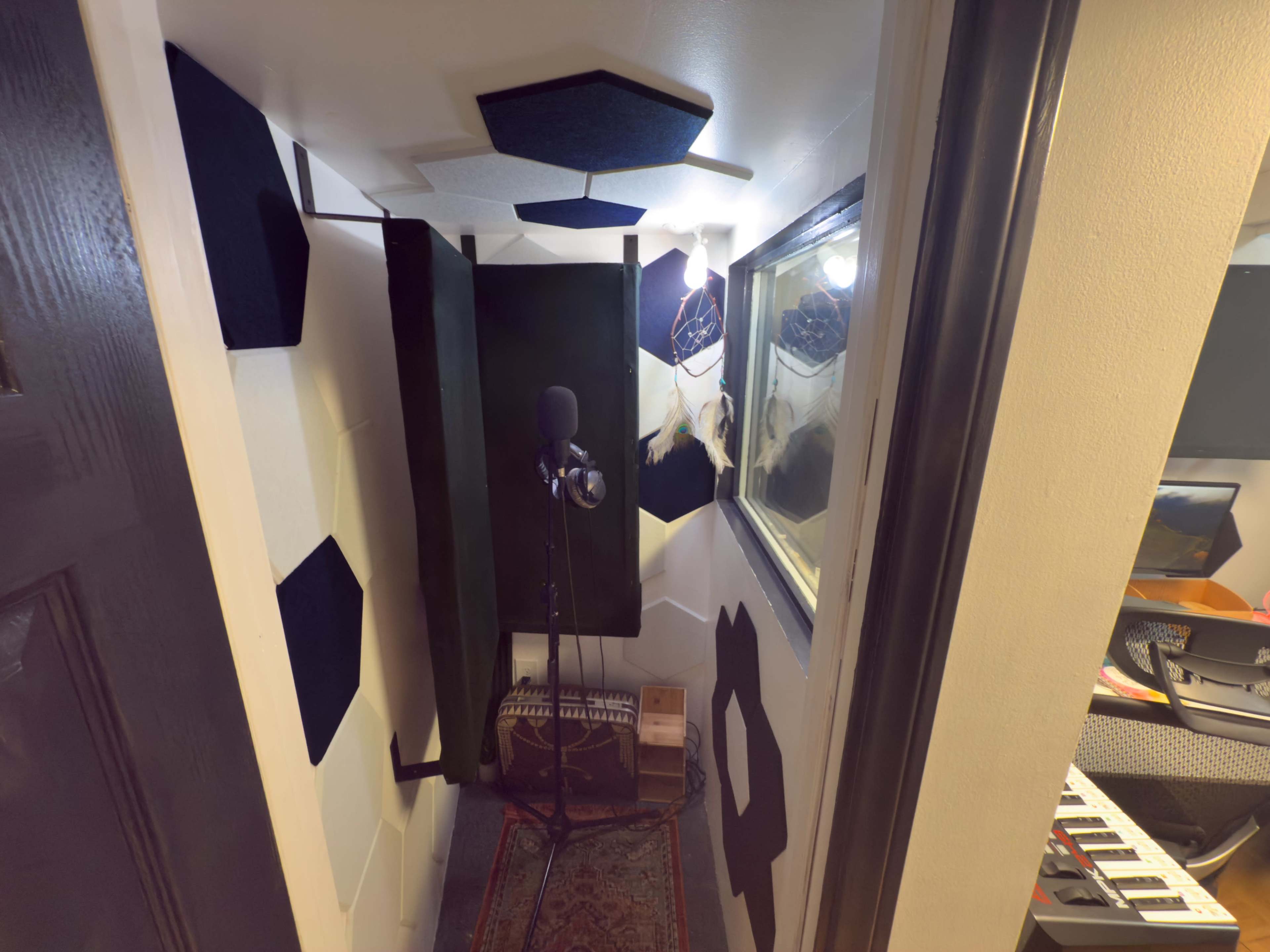 The image shows a small recording booth with acoustic panels on the walls, a microphone stand in the center, and a window overlooking another room with musical instruments.