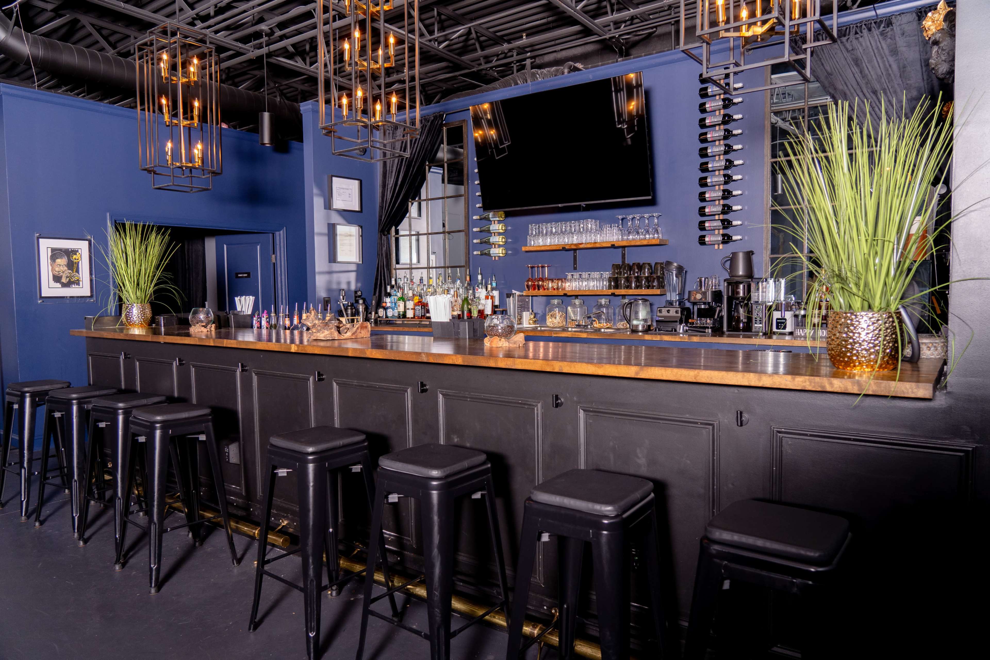 A modern bar with a long wooden countertop, black stools, and shelves displaying various bottles and glasses against a blue wall.
