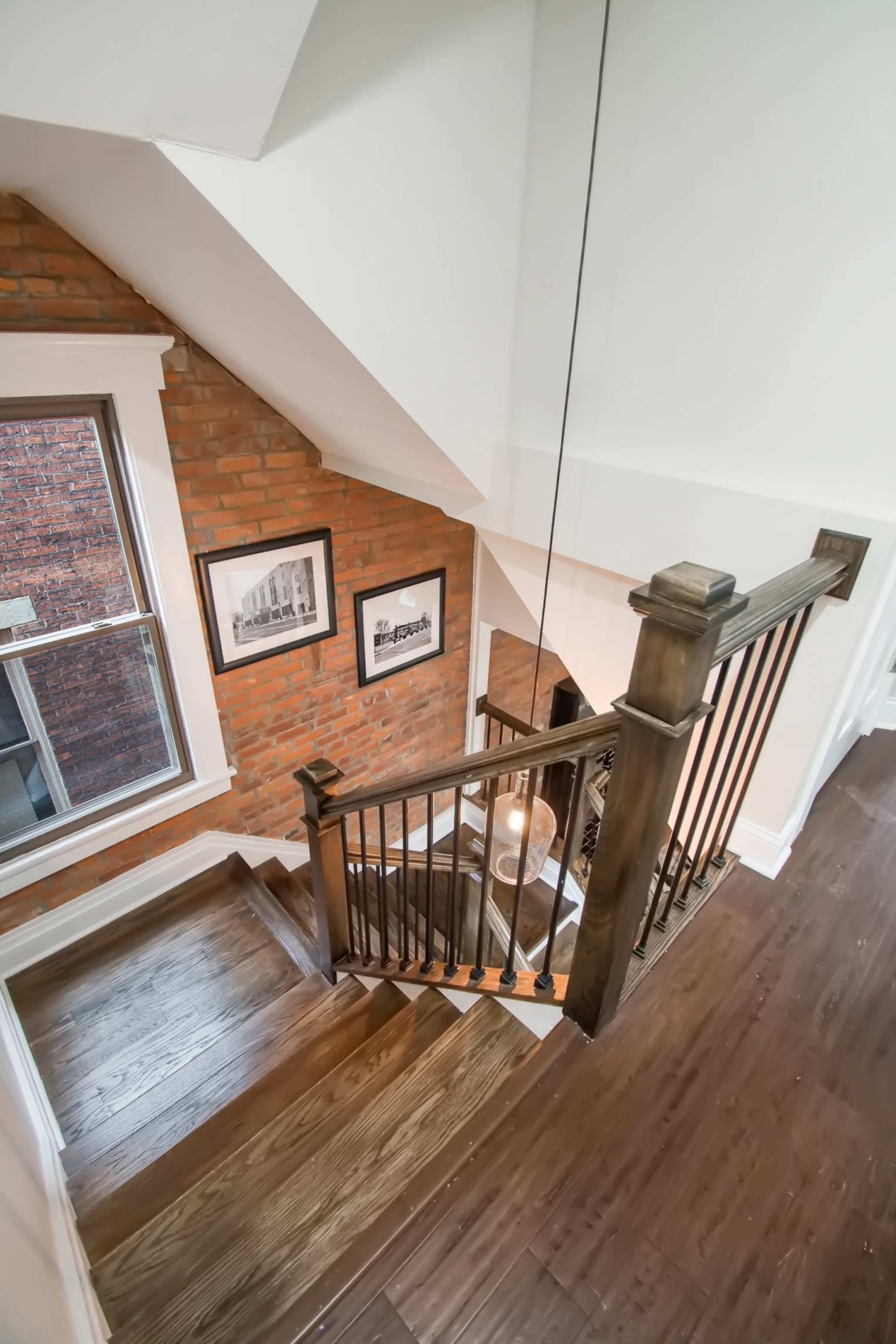 A staircase with wooden railings descends past a brick wall, leading to a well-lit landing with framed photographs and a window.