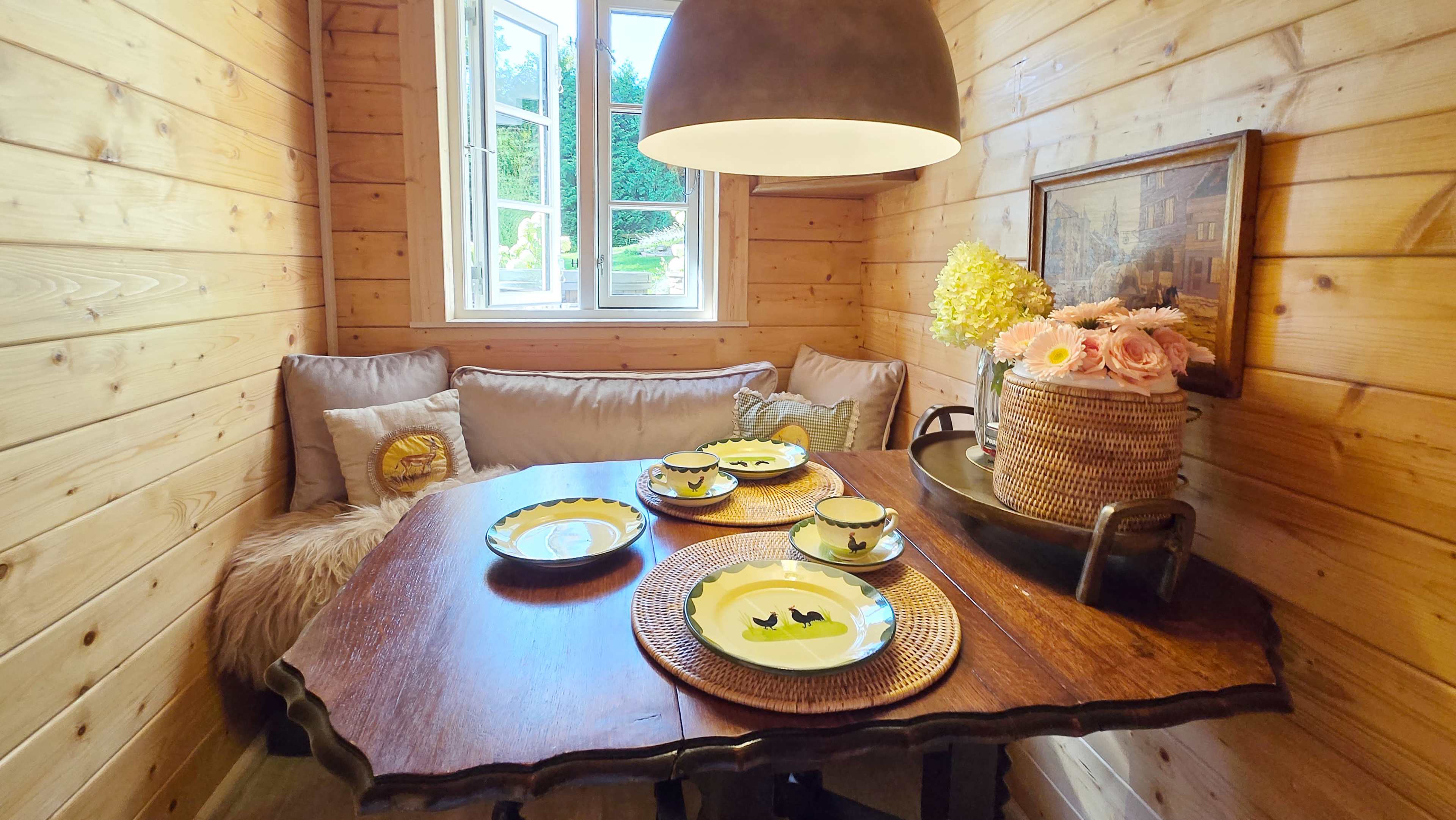 A cozy, wooden dining nook features a round table set with yellow plates and decorative items, illuminated by a large pendant light.