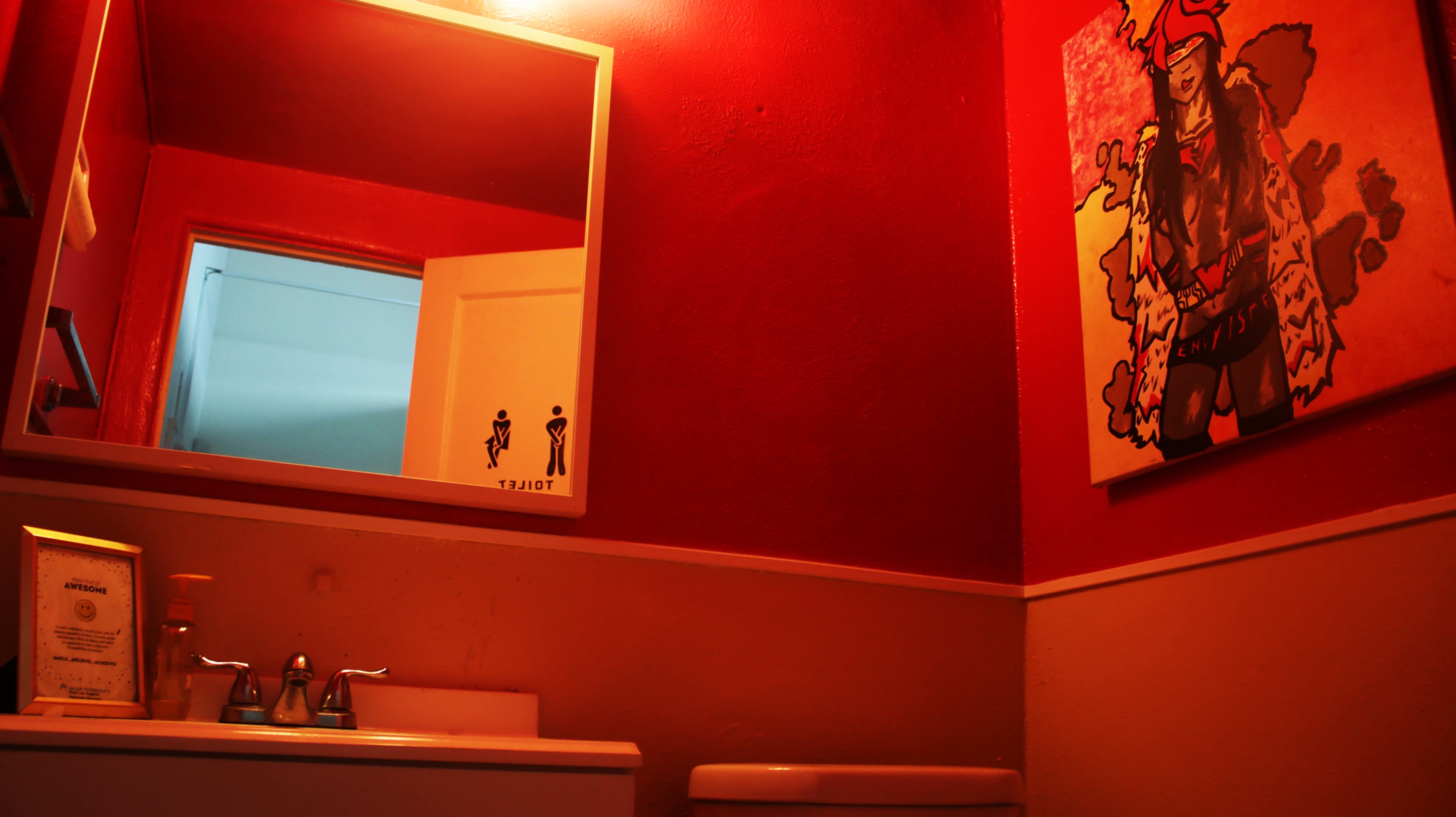 The image shows a small bathroom with red walls, a mirror reflecting a door, a sink, and a toilet, accompanied by a colorful artwork on the wall.