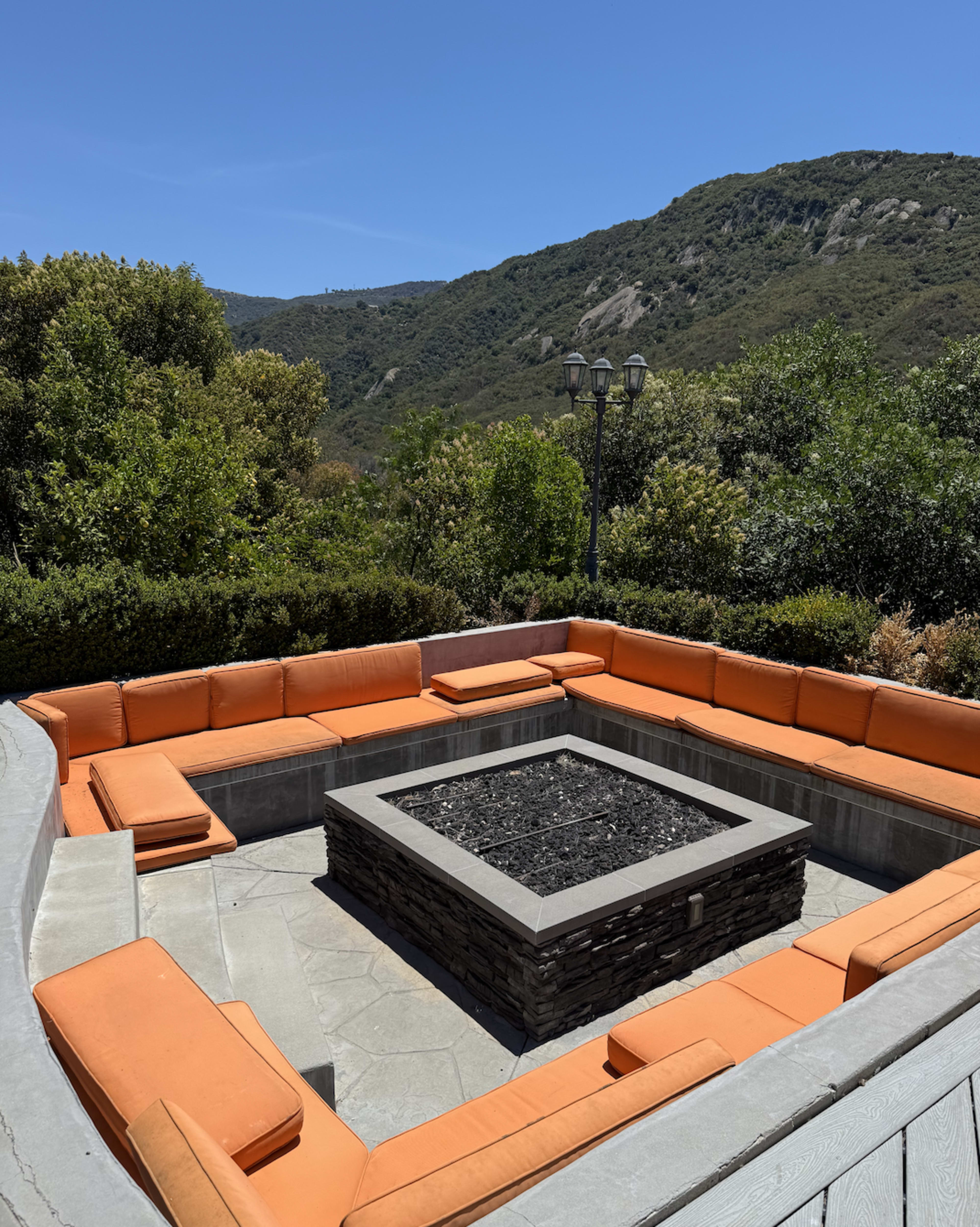 The image shows a square seating area with orange cushions surrounding a fire pit, set against a backdrop of green hills under a clear blue sky.