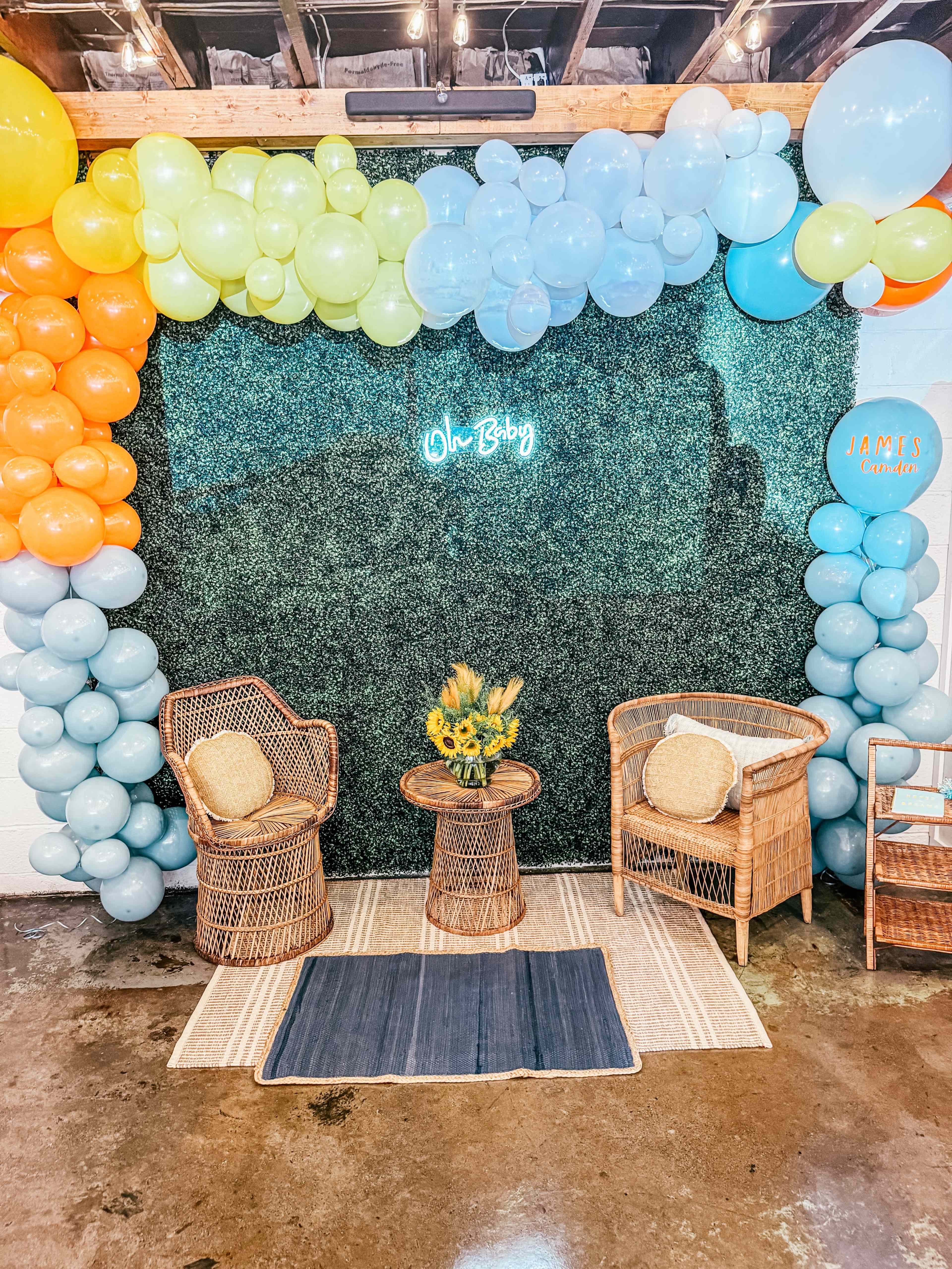 A cozy setup featuring two rattan chairs and a circular table against a backdrop of colorful balloons and greenery, with a neon sign that reads "Oh Baby."