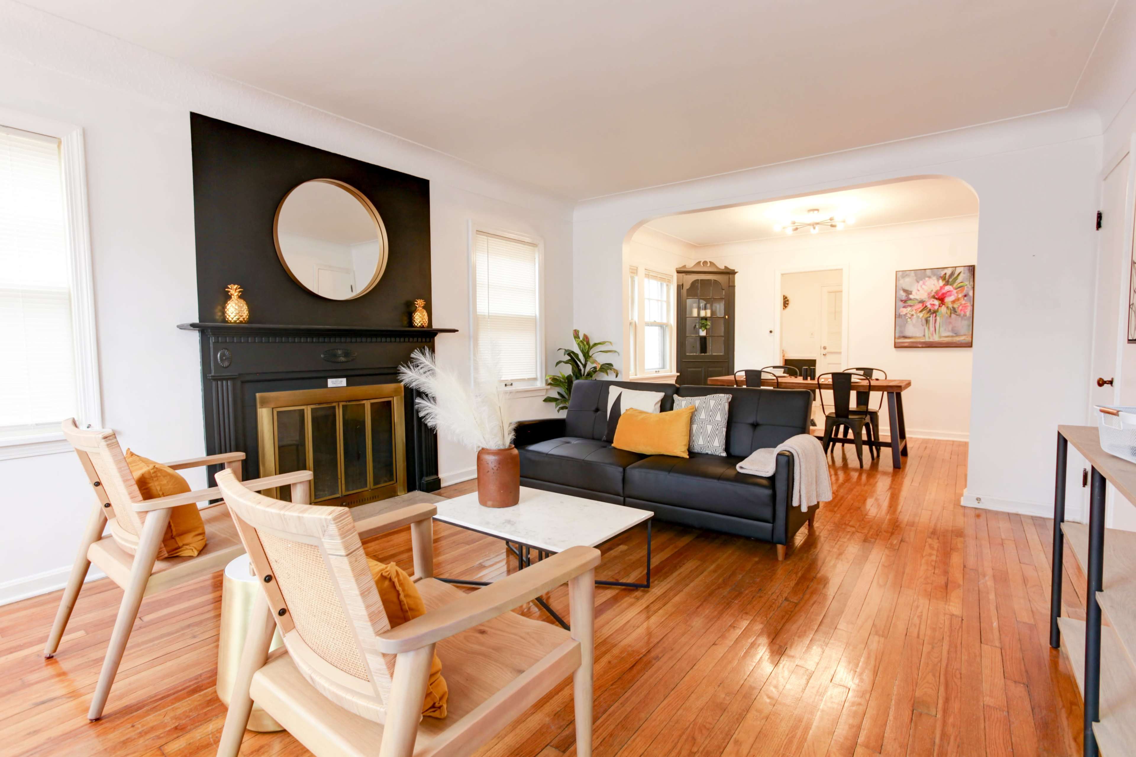 A living room features a black fireplace with a round mirror above it, a sofa with an orange accent pillow, wooden chairs, and a dining area visible in the background.