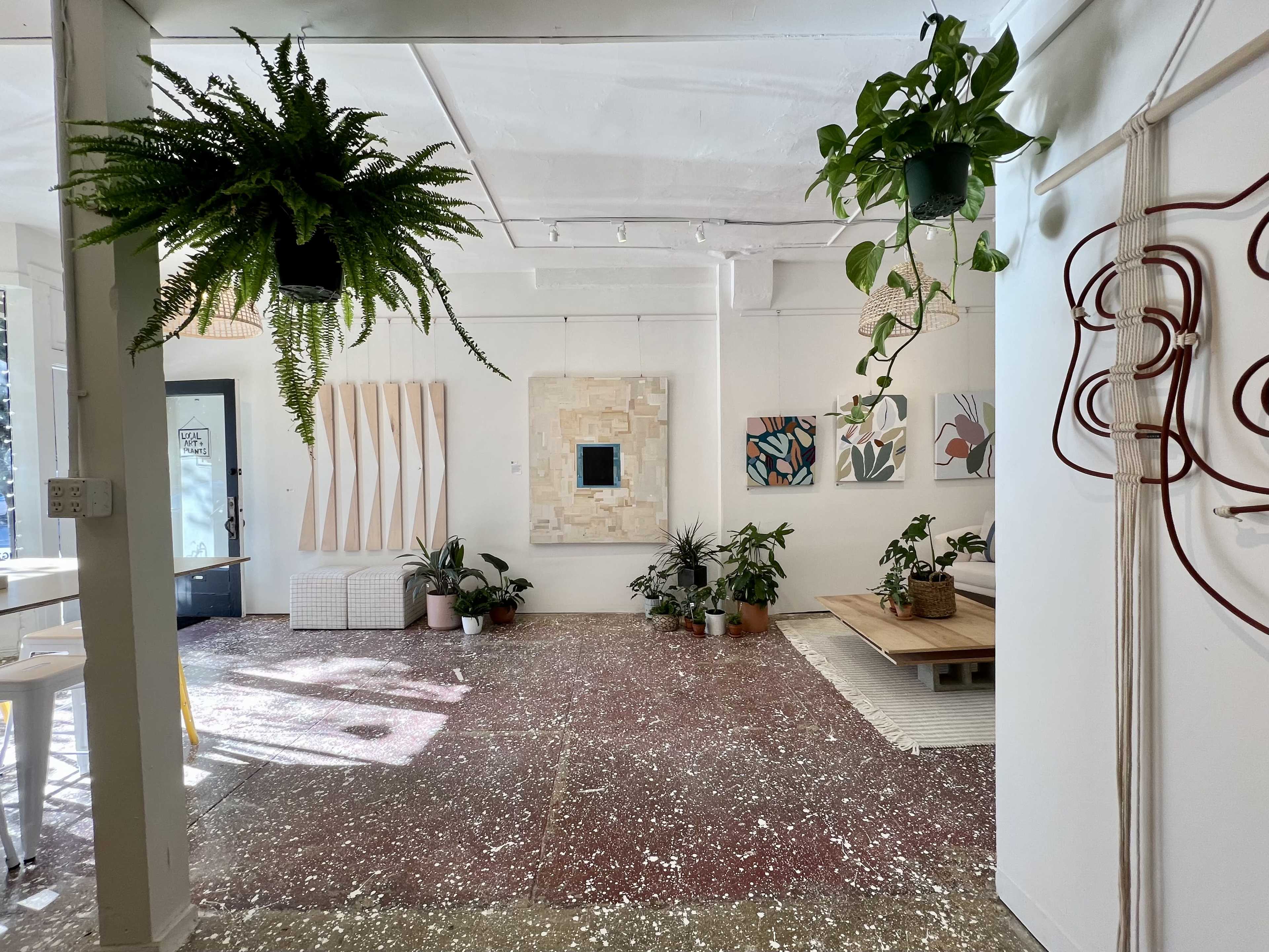 A brightly lit contemporary interior space featuring plants, artwork on the walls, and a mixture of furniture and decorative elements on the floor.