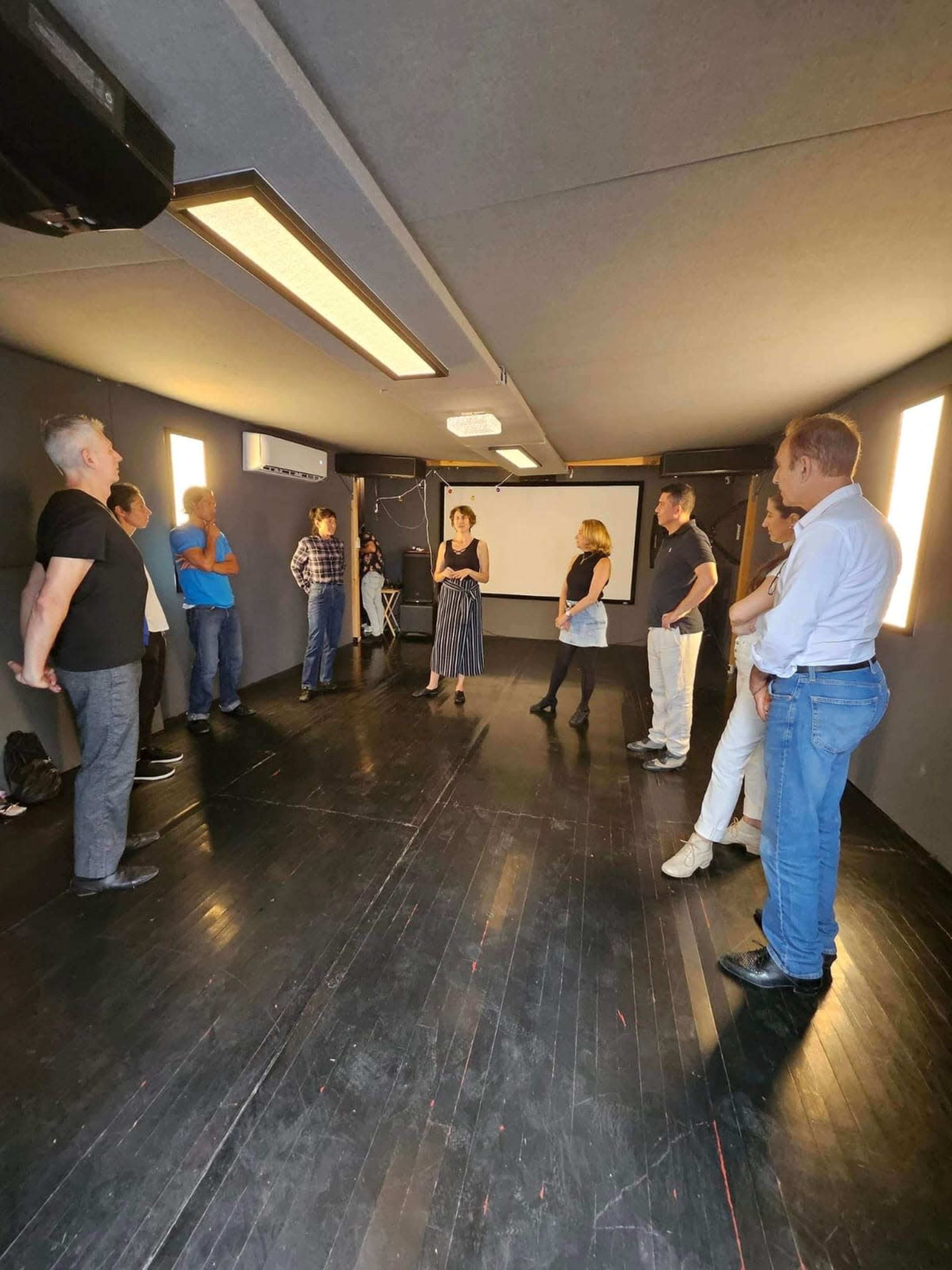 A group of people stands in a dimly lit room with black floors and walls, facing a woman who appears to be speaking or leading a discussion.
