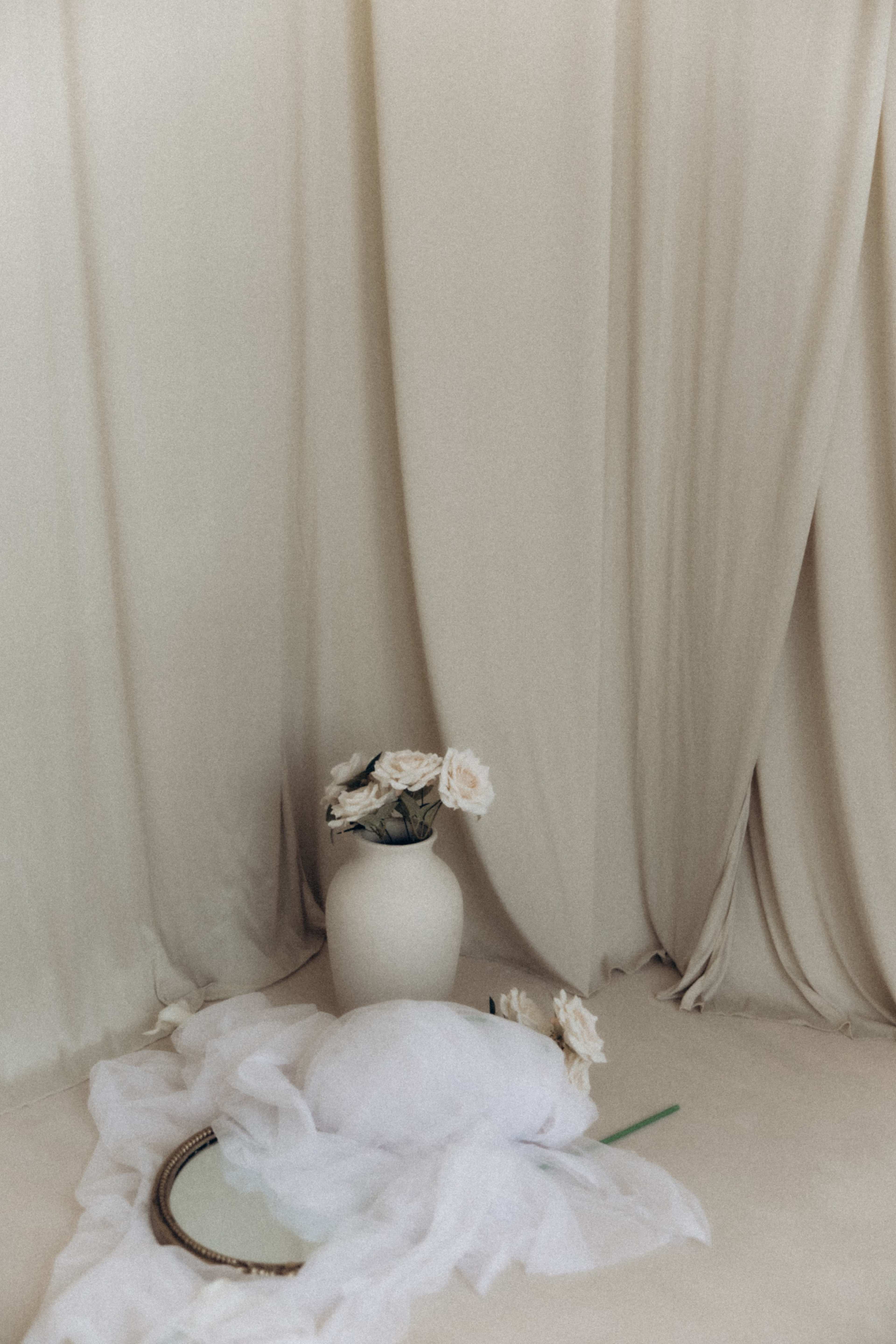A white vase with flowers sits on a light surface in front of draped beige fabric.