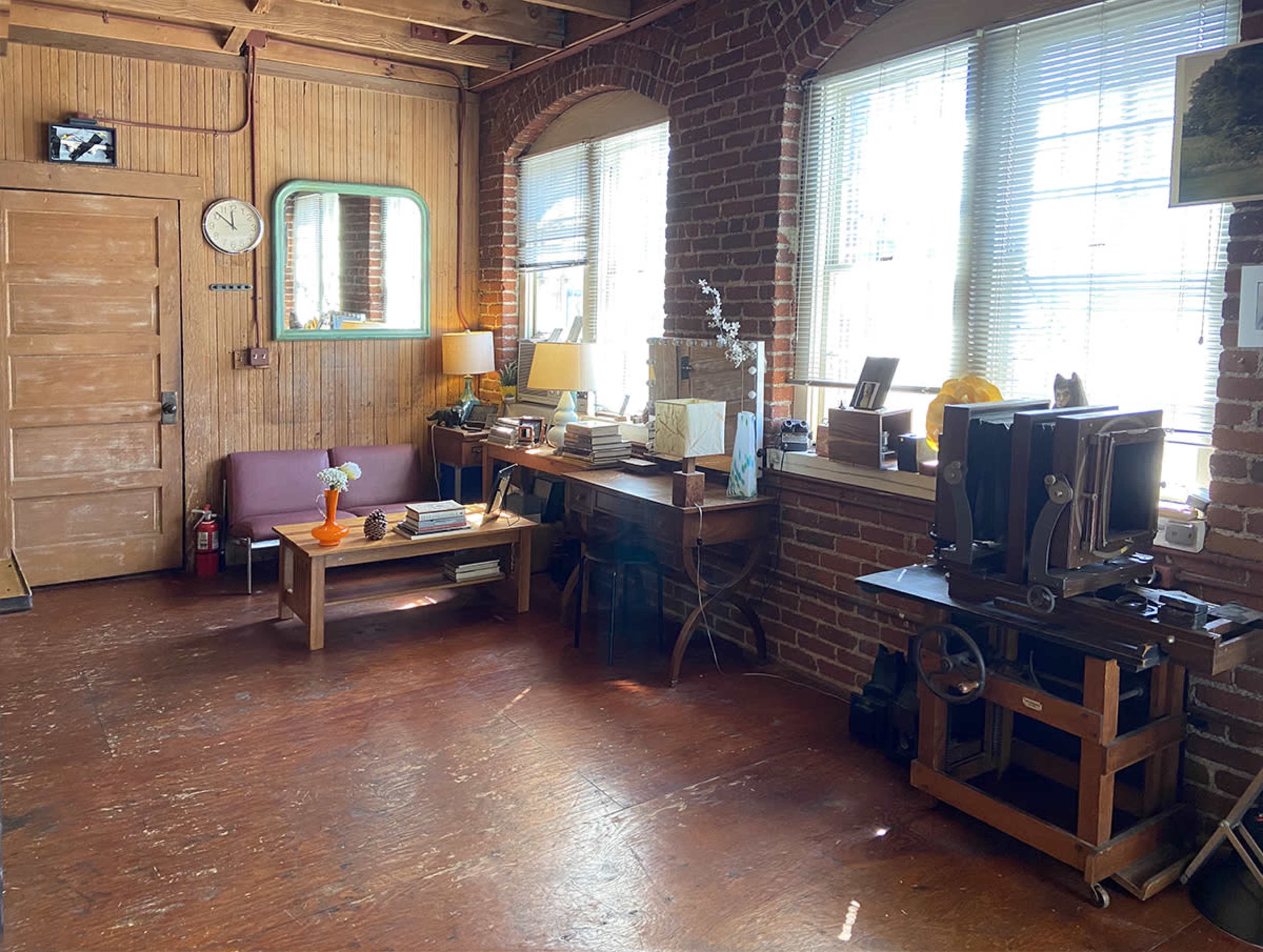 A workspace featuring wooden furniture, large windows, and exposed brick walls, with a clock, lamp, and various books and objects arranged on the desk.