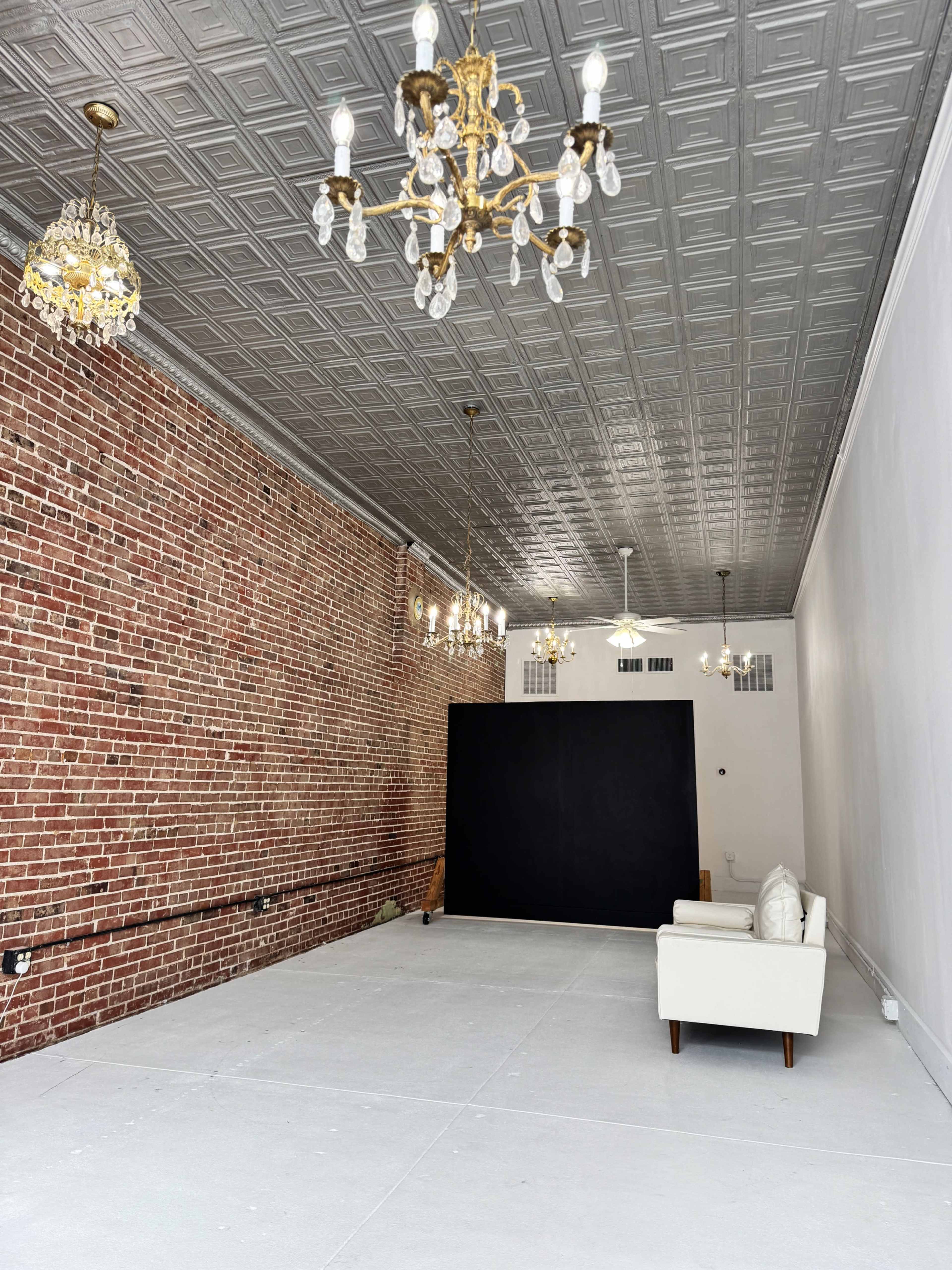 The image shows an empty interior space featuring exposed brick walls, a smooth white floor, and several decorative chandeliers hanging from a tin ceiling, with a black backdrop and a white sofa positioned along one side.