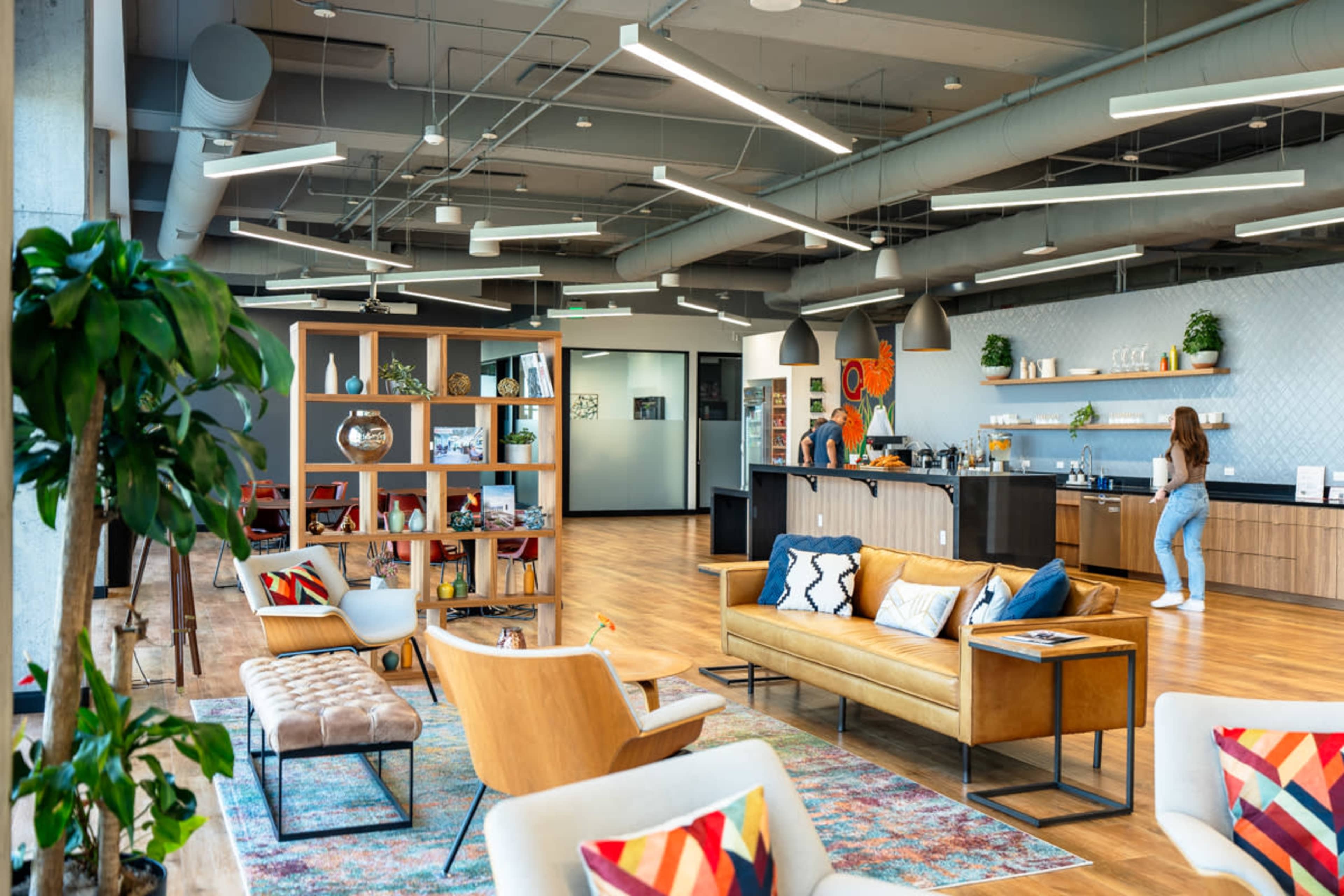 The image shows a modern office lounge area with comfortable seating, a bookshelf, and a kitchen counter, featuring wooden floors and ample natural light.