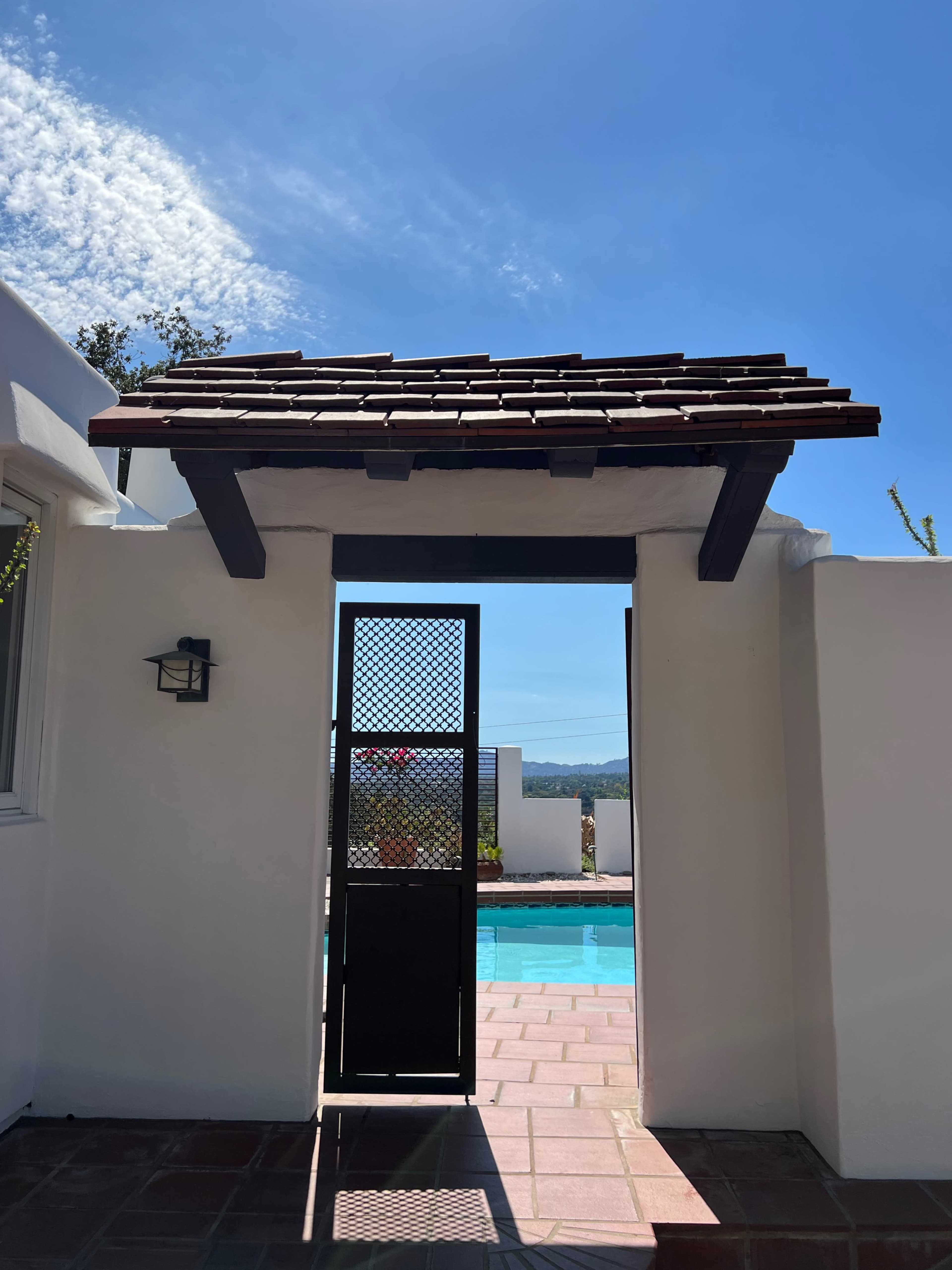 A black gate with a wooden awning frames a view of a blue pool and distant hills under a clear sky.
