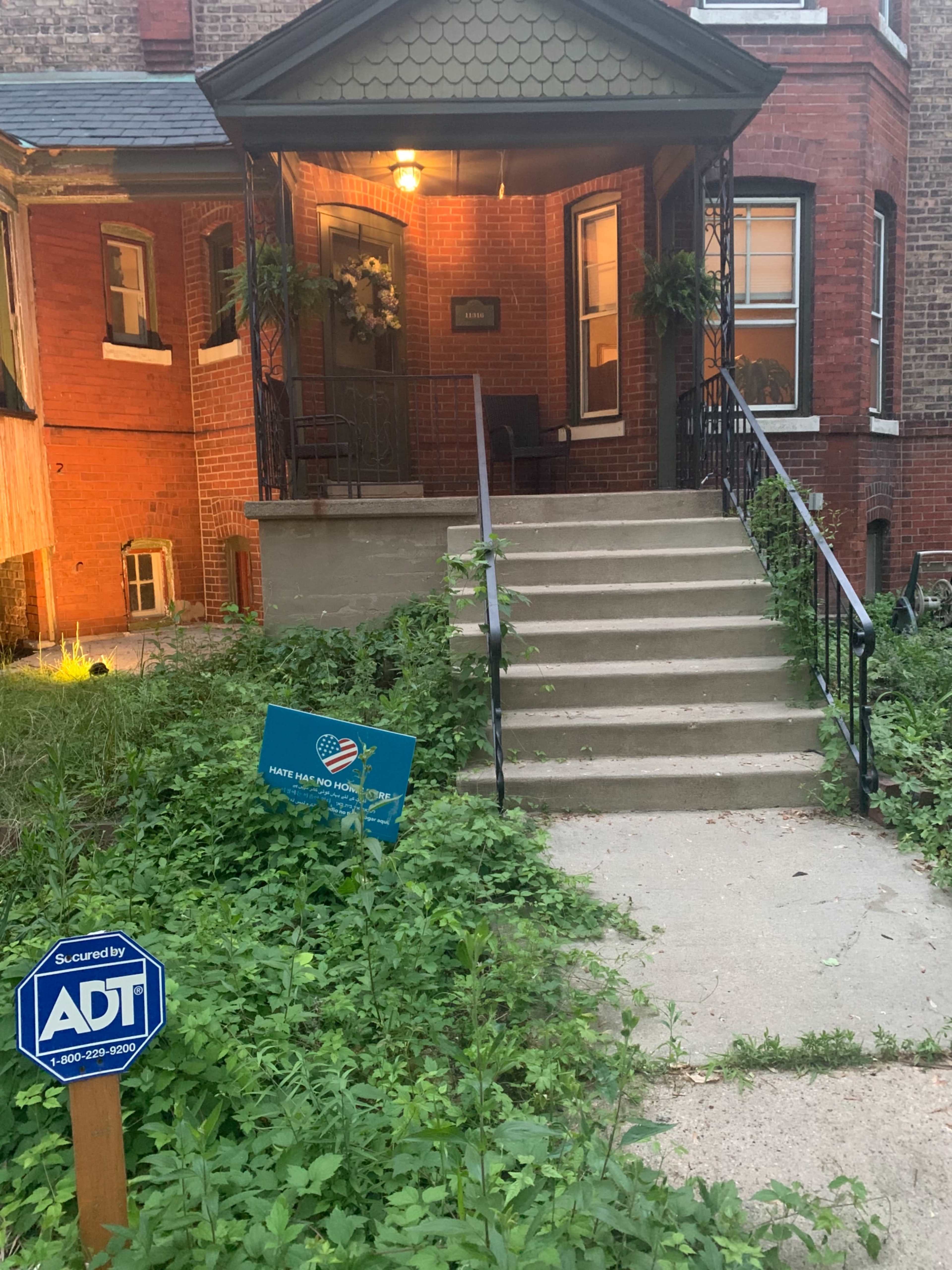 A residential entrance with a set of concrete steps leading to a porch, surrounded by overgrown grass and plants, with an ADT security sign positioned on the ground.