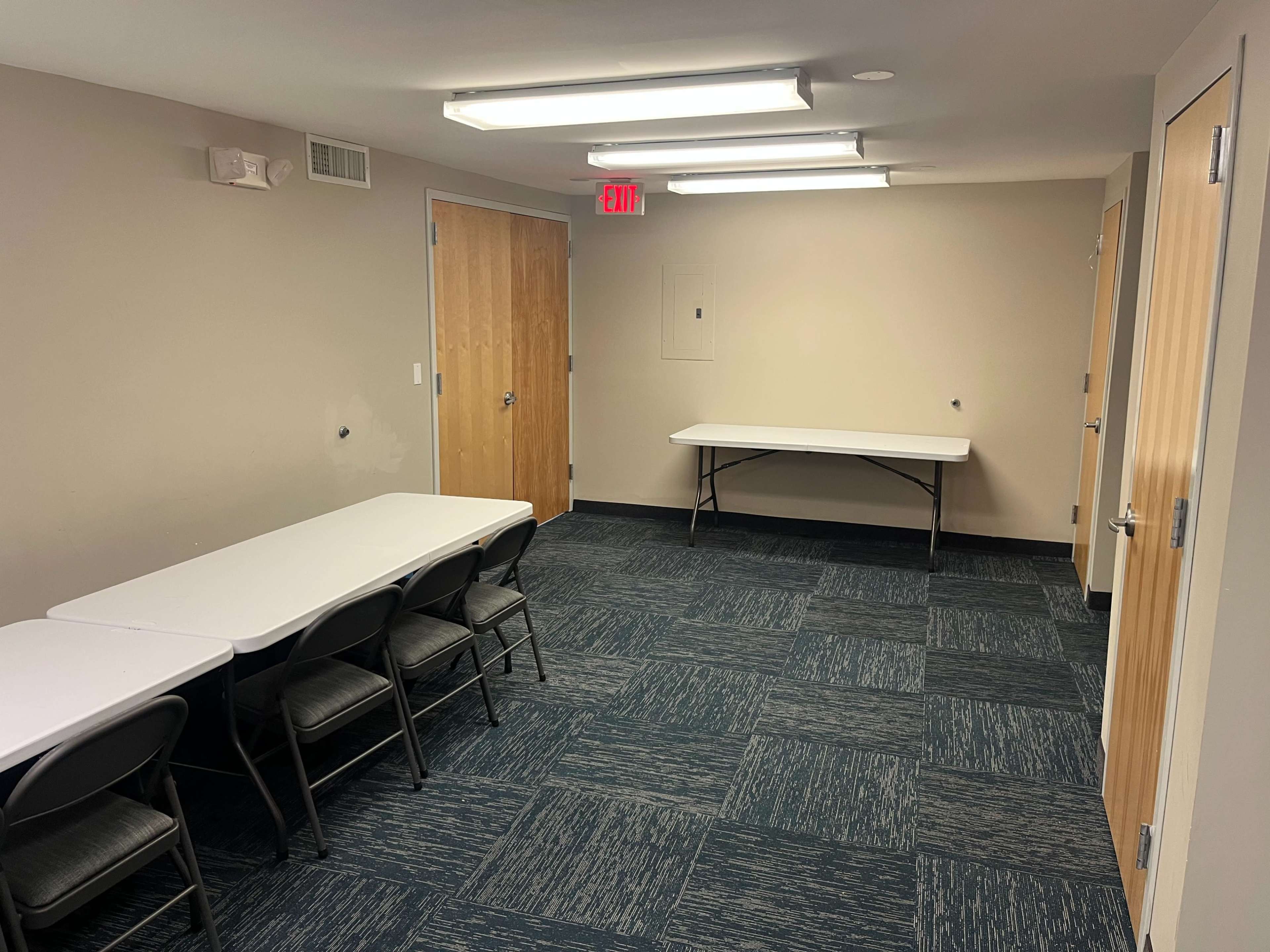 A small empty room with a table and several chairs arranged at one end, and two doors on opposing walls.