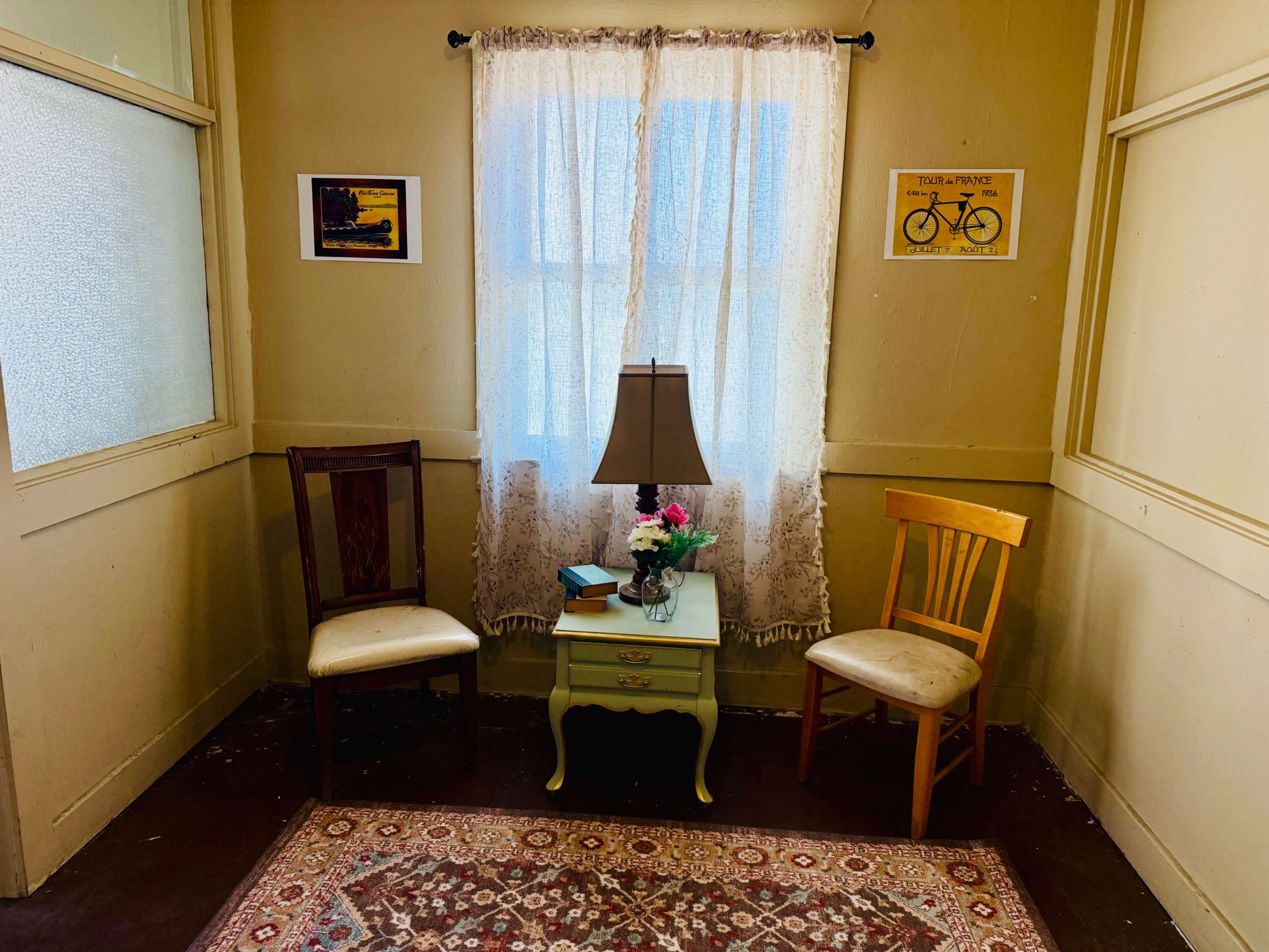 The image shows a small corner of a room with two chairs, a side table with a lamp and flowers, and a window covered by a sheer curtain.