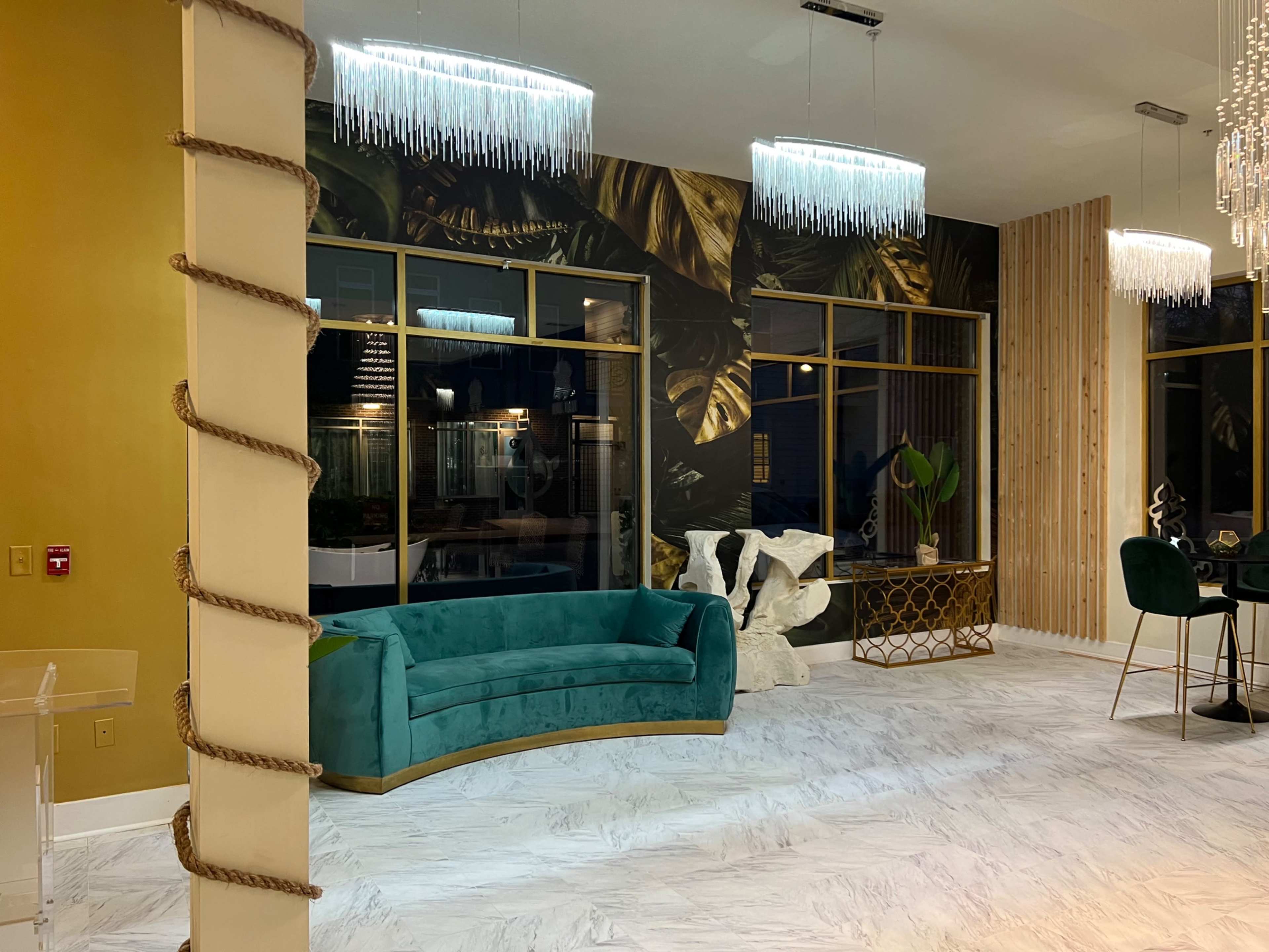 A modern interior space with a teal sofa, decorative wall art featuring tropical leaves, and various stylish furnishings including a unique sculpture and a dining table.